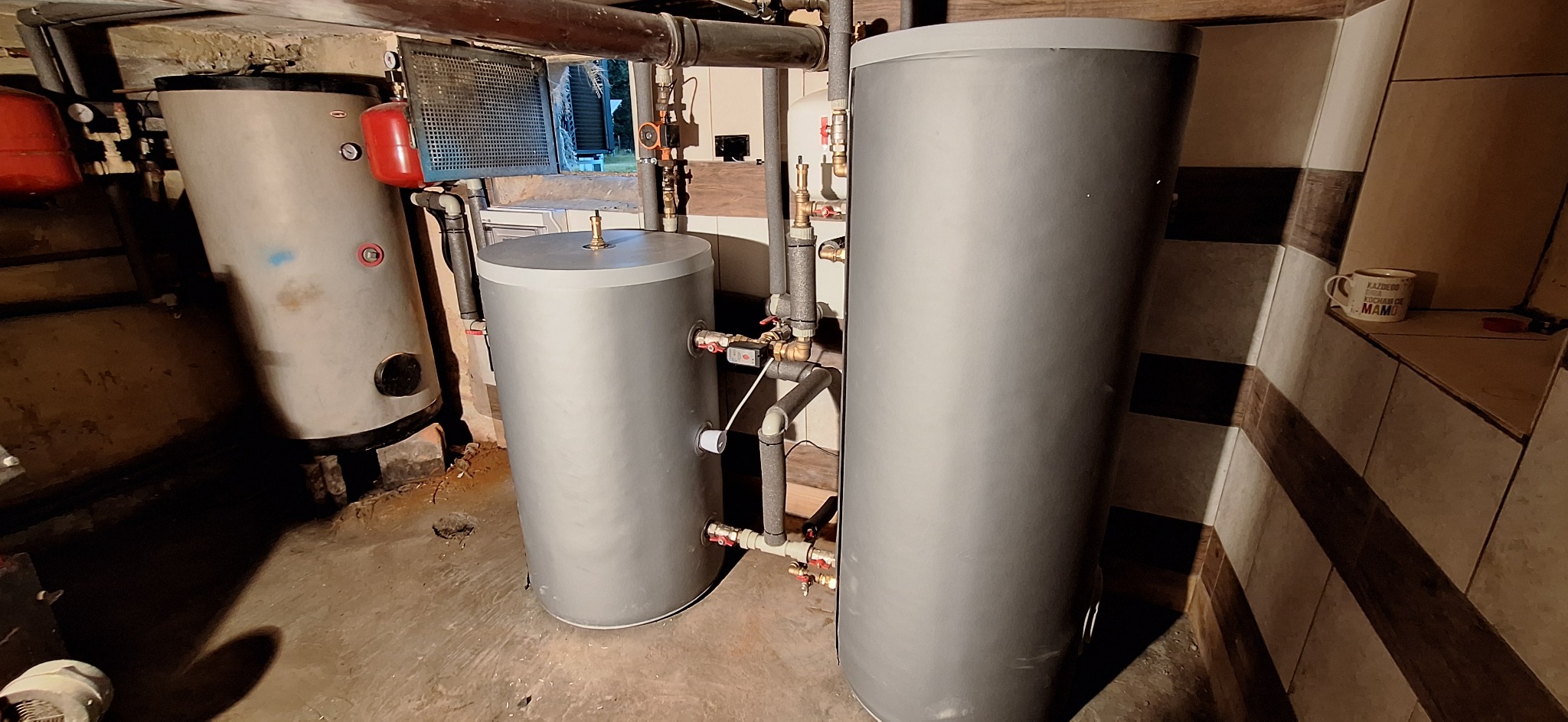 Trzy szare zbiorniki na wodę różnej wielkości w piwnicy, z widocznymi rurami, zaworami i grzejnikiem, oraz kubek z napisem na półce w tle.
