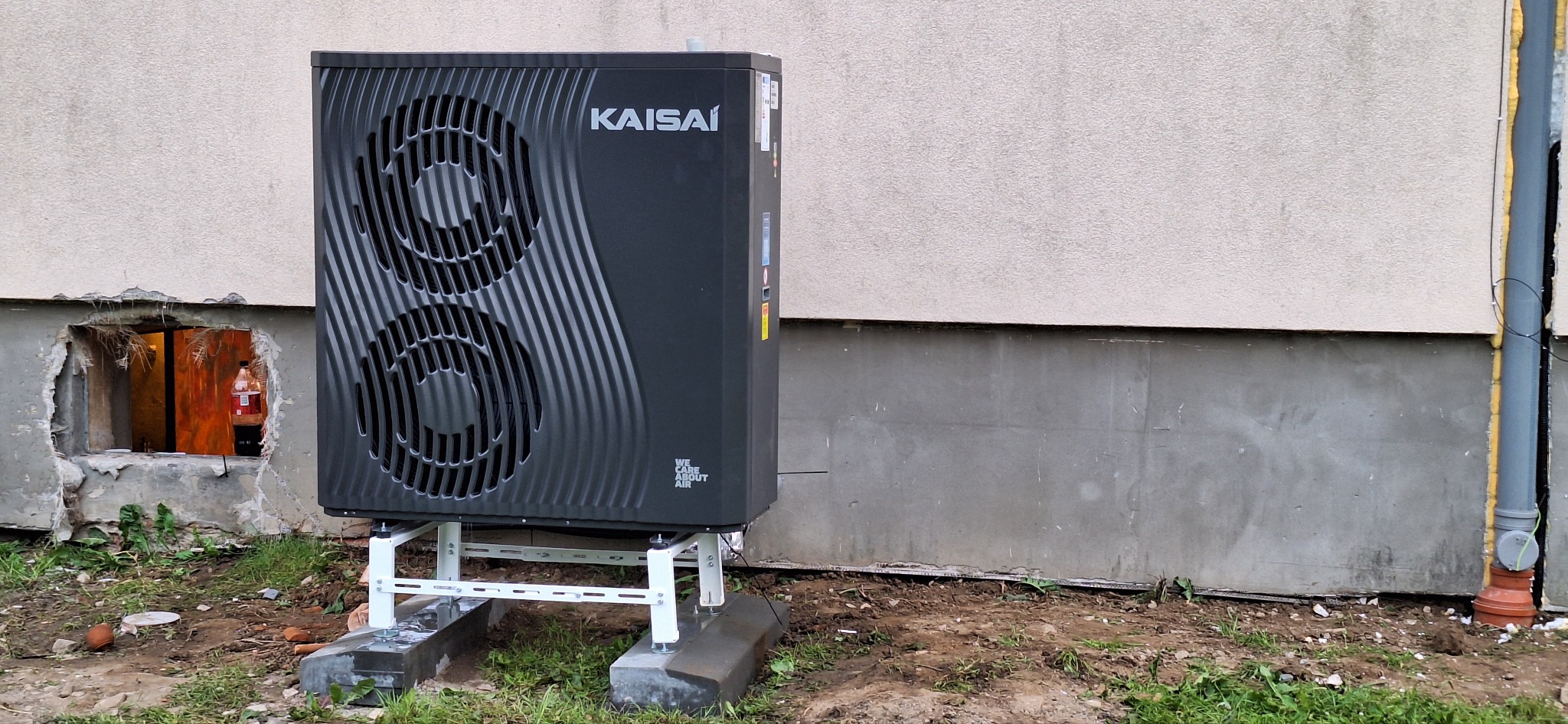Czarna pompa ciepła marki Kaisai zamontowana na białym stelażu przed elewacją budynku z widocznym otworem w fundamencie i butelką w środku.