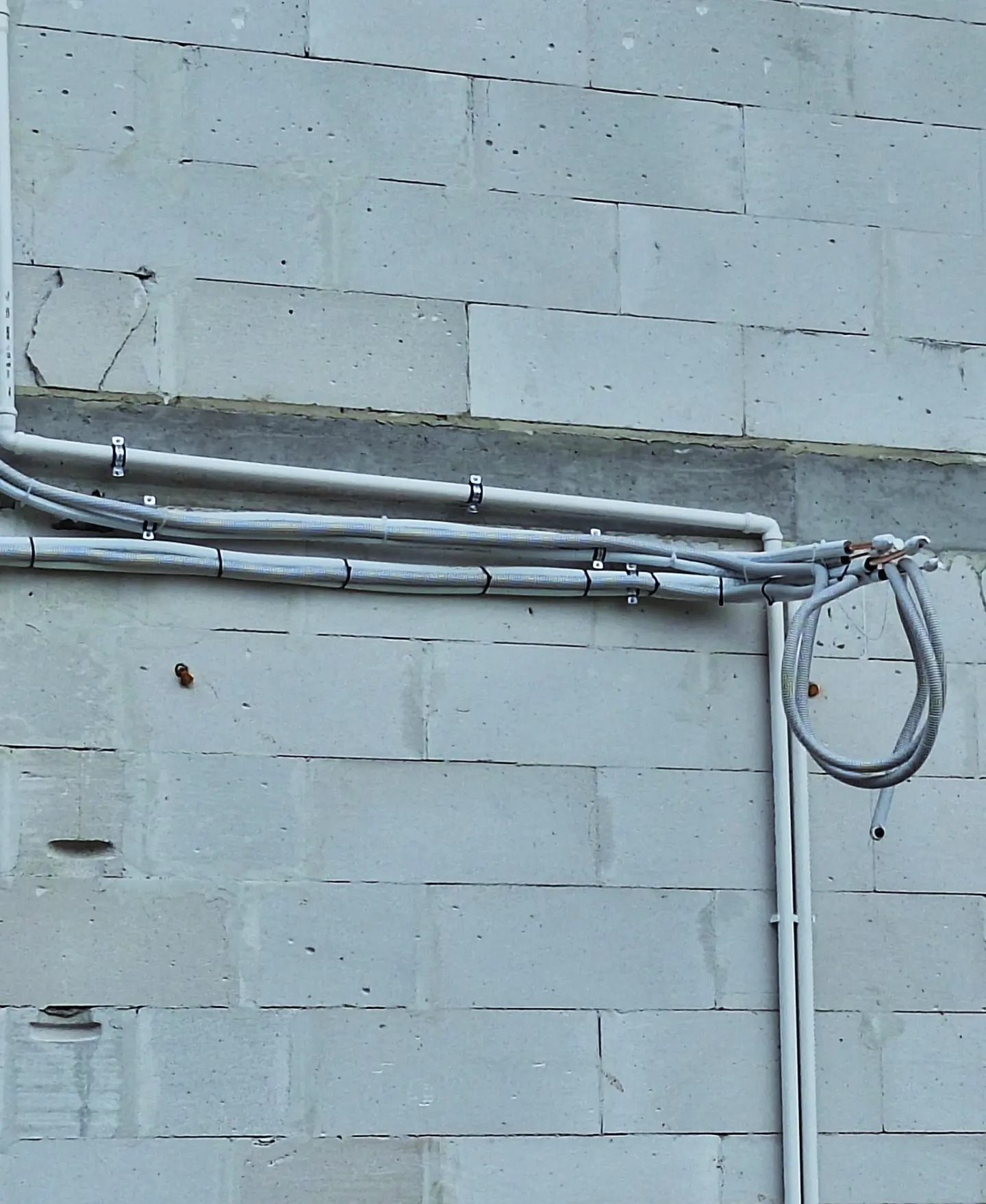 Szare rury w izolacji, przytwierdzone metalowymi uchwytami do ściany z białych bloczków betonowych. Pionowa rura z tworzywa sztucznego biegnie wzdłuż ściany.