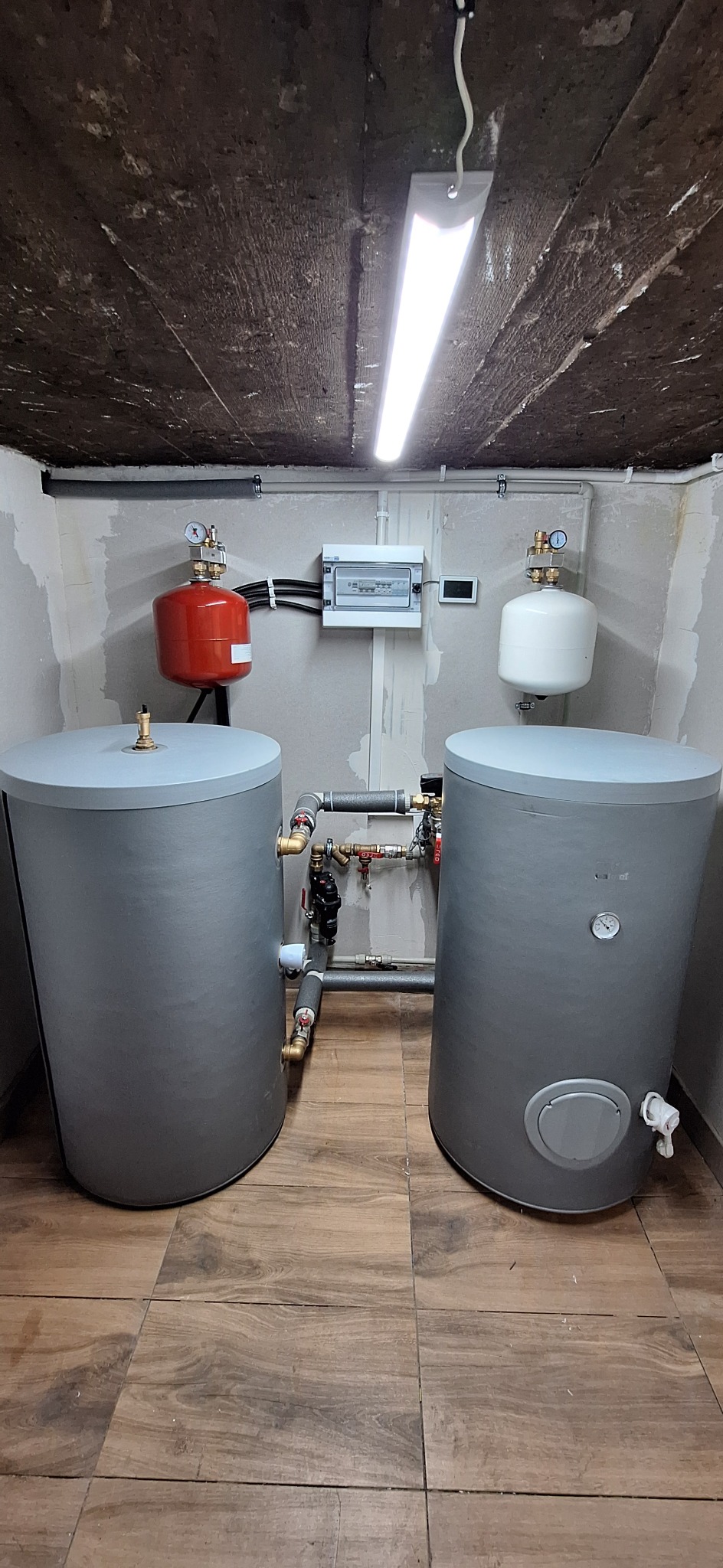Dwa szare zbiorniki akumulacyjne połączone rurami w pomieszczeniu z drewnopodobną podłogą, widoczne również czerwony i biały zbiornik wyrównawczy, skrzynka elektryczna i surowy betonowy sufit z lampą.