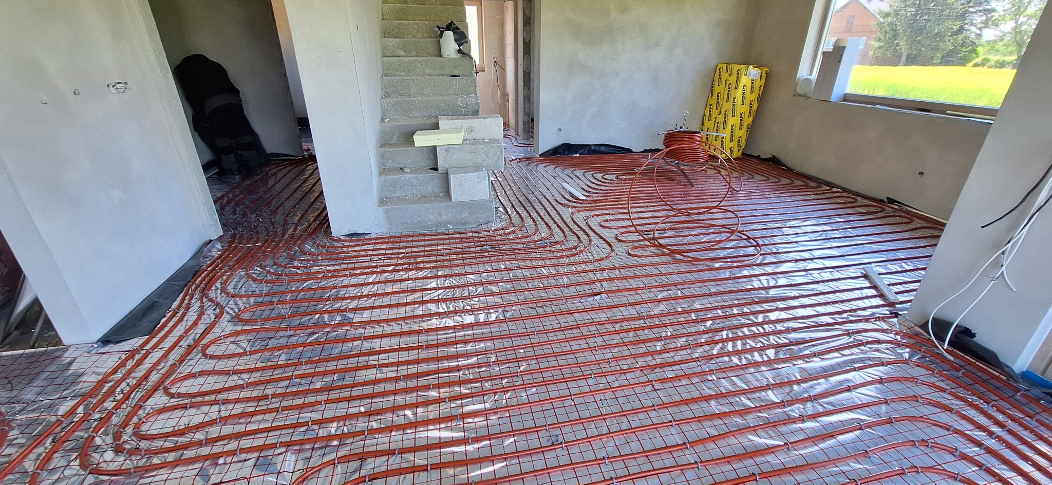 Instalacja ogrzewania podłogowego: rury grzewcze ułożone na folii odblaskowej i siatce, widoczne elementy konstrukcyjne budynku w stanie surowym.