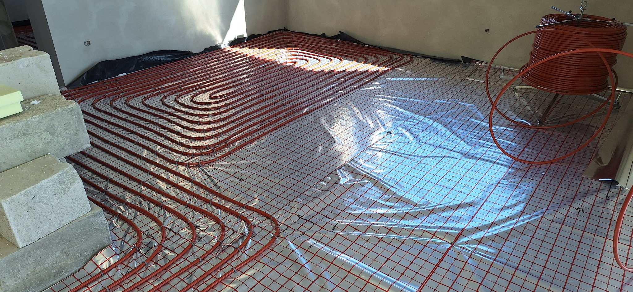 Czerwone rury ogrzewania podłogowego ułożone na folii z siatką w surowym wnętrzu, z blokami betonowymi w rogu i zwiniętymi rurami w tle.