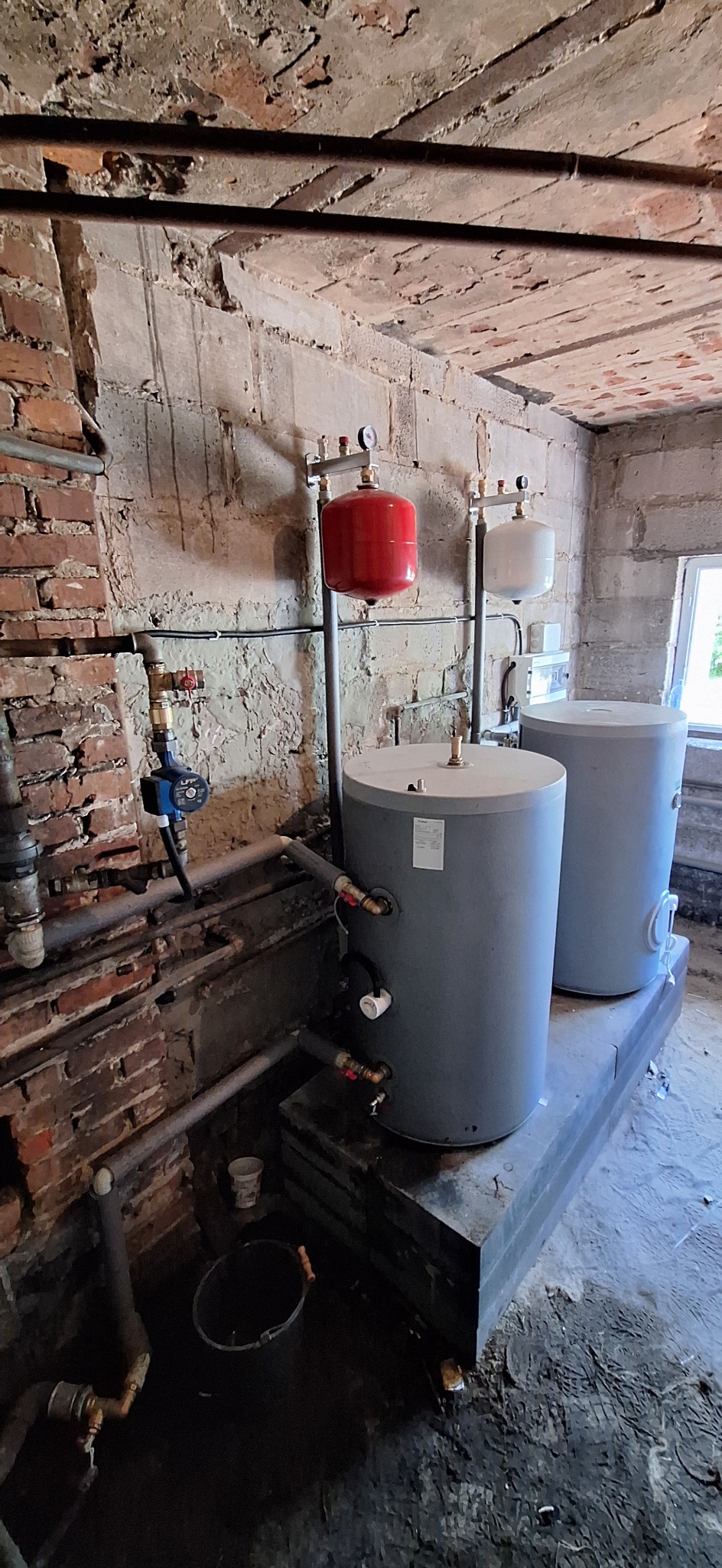 Dwa szare zasobniki ciepłej wody stojące na betonowej podstawie w pomieszczeniu z ceglanymi ścianami i widocznymi rurami, czerwony zbiornik wyrównawczy na ścianie, widoczny fragment okna.