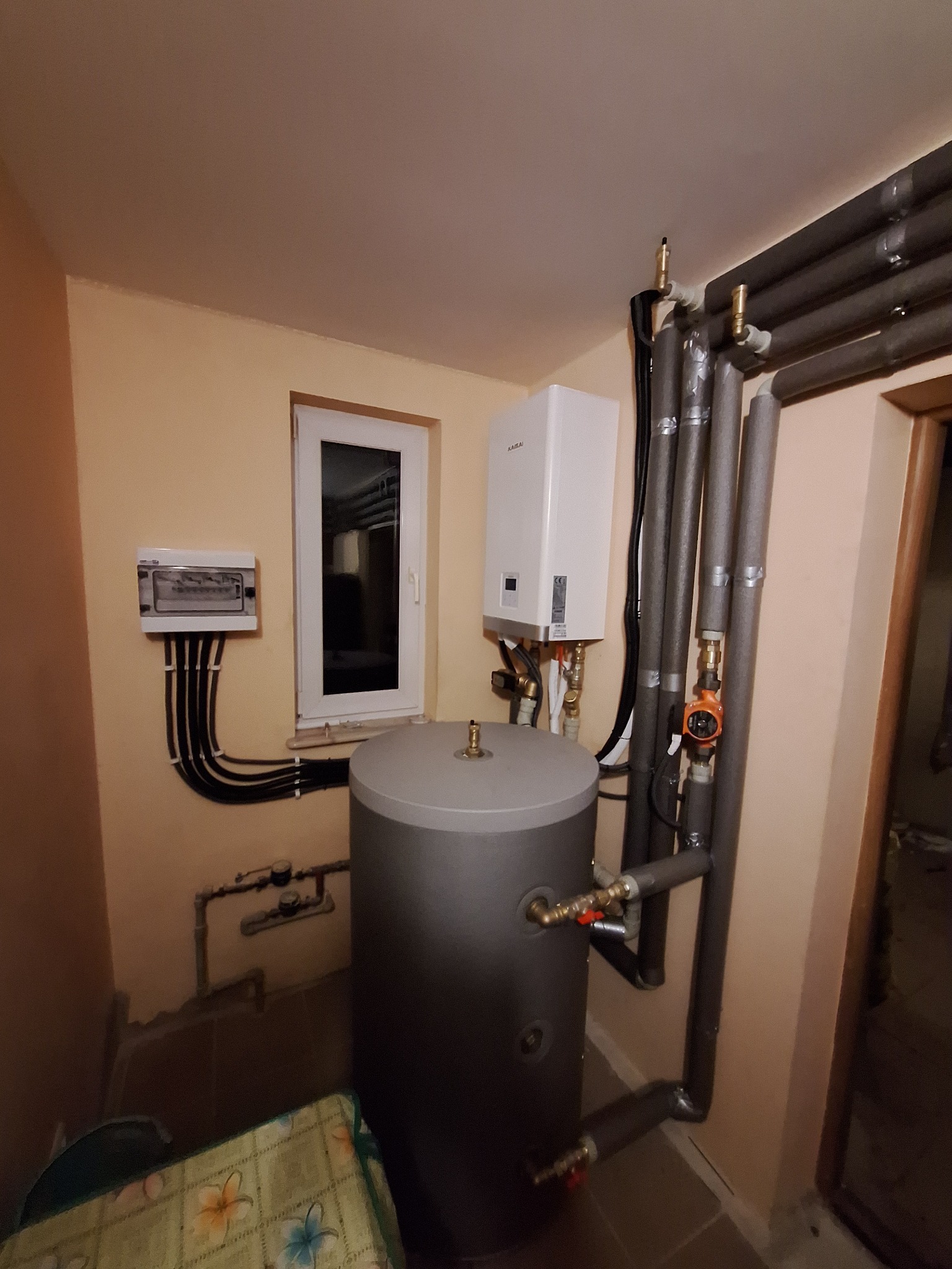 Kotłownia w piwnicy: szary zbiornik akumulacyjny, biały piec gazowy Kaisai, rury w izolacji termicznej z pompką obiegową, oraz skrzynka elektryczna z czarnymi przewodami przy oknie.