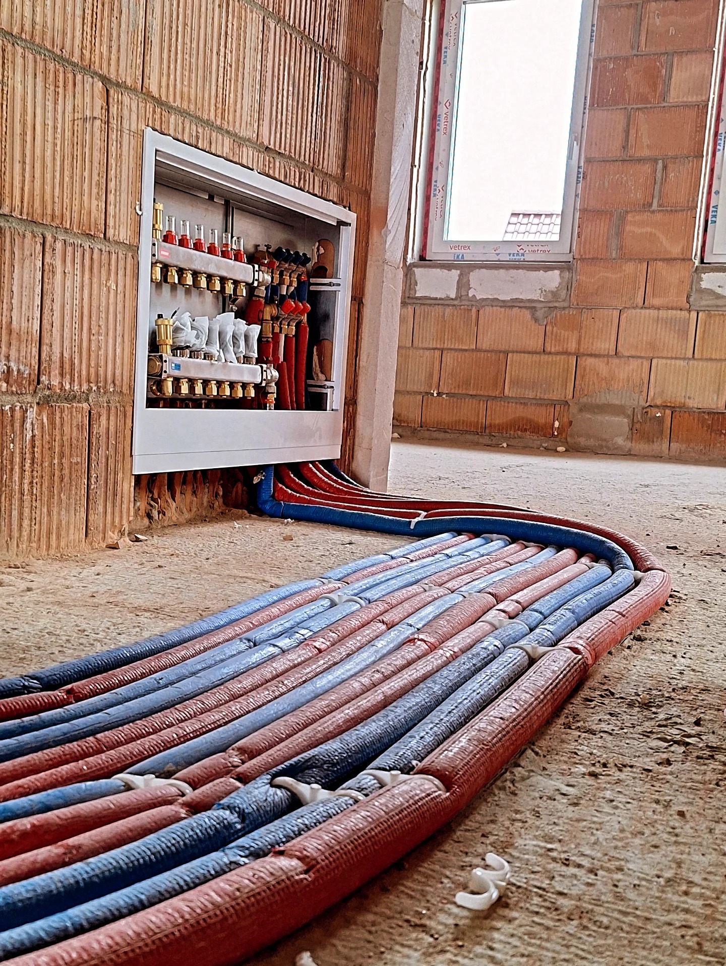 Rozdzielacz ogrzewania podłogowego z rurami w kolorze czerwonym i niebieskim, ułożonymi w pętle na podłodze w surowym stanie, widok z niskiej perspektywy.