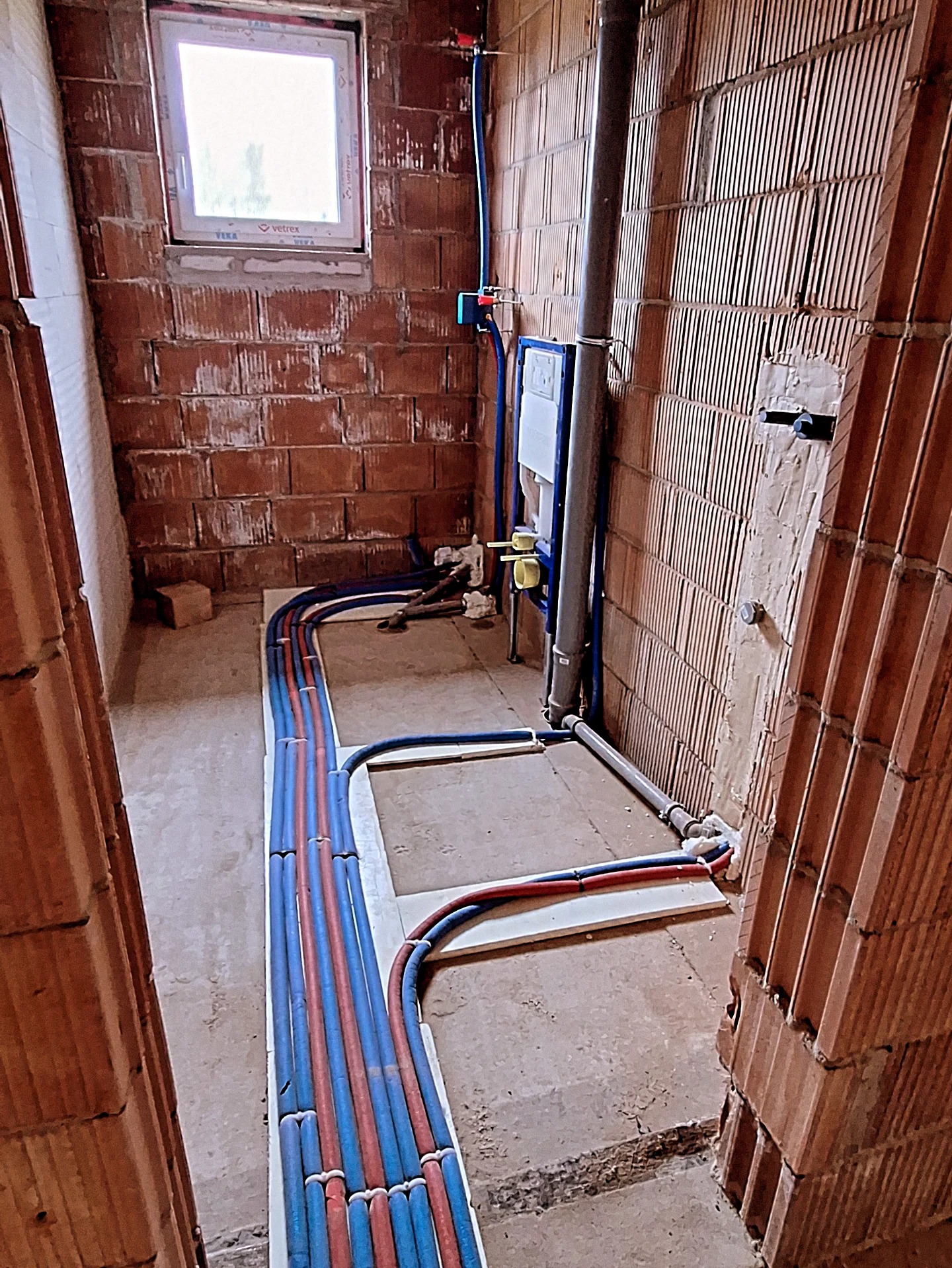 Instalacja wodna i kanalizacyjna w surowej łazience z cegły. Widoczne rury w podłodze i przyłącza do toalety podtynkowej. Okno w tle.