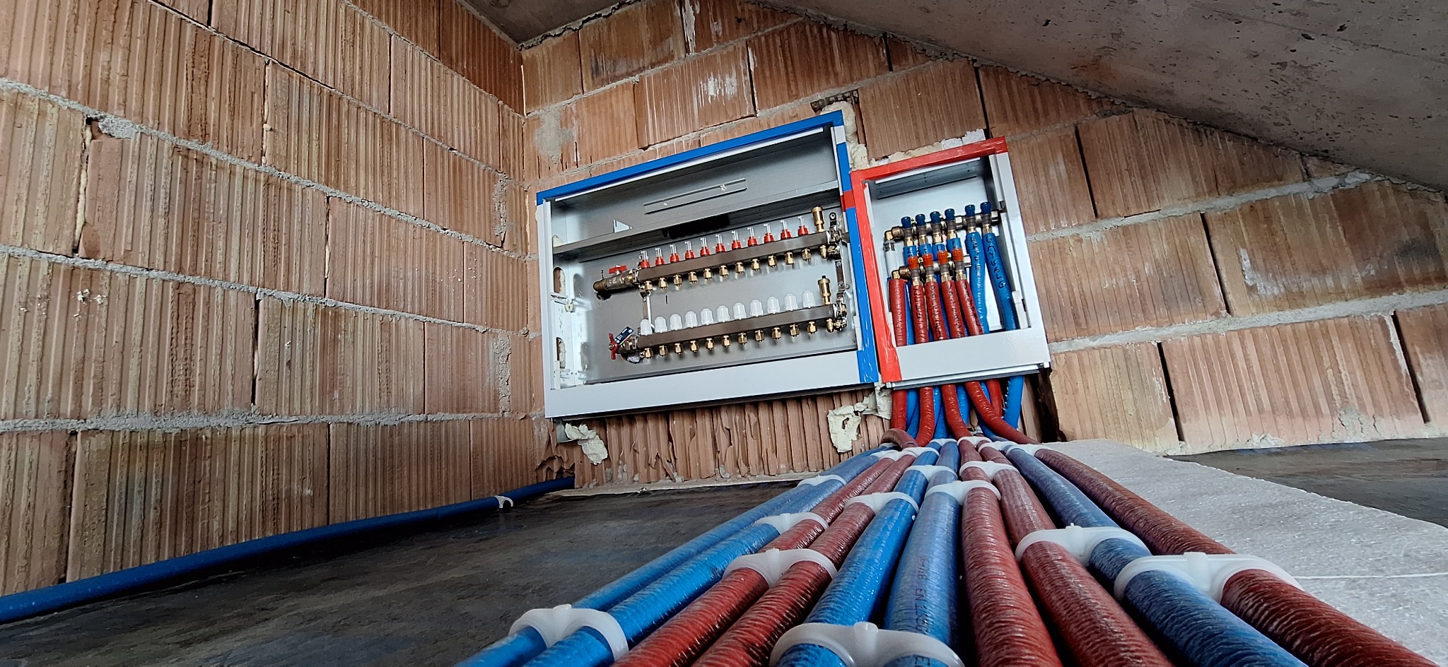 Rozdzielacz ogrzewania podłogowego z rurami w kolorze czerwonym i niebieskim, zamontowany na ścianie z cegły dziurawki w surowym stanie. Widok z dołu.