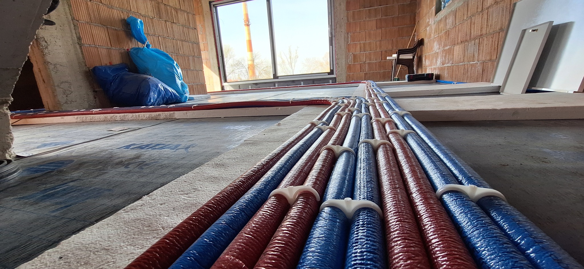 Ułożone rury ogrzewania podłogowego w kolorze czerwonym i niebieskim, przymocowane białymi uchwytami na izolacji, widok z niskiej perspektywy w surowym wnętrzu.