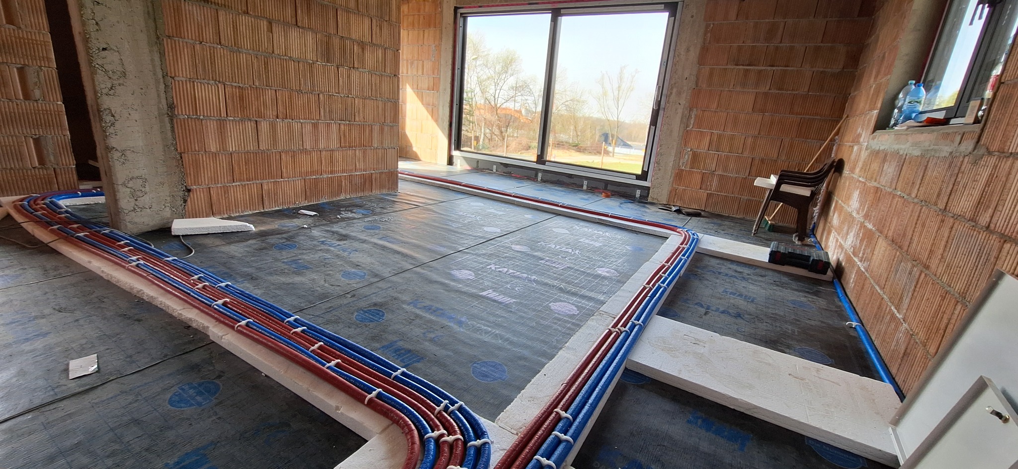 Układanie rur ogrzewania podłogowego na izolacji w surowym wnętrzu. Czerwone i niebieskie rury przymocowane do styropianu. Widok na okno i ceglane ściany.