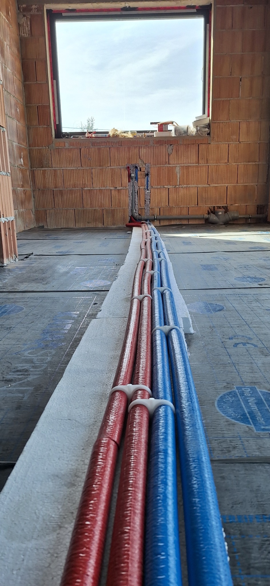Instalacja rur grzewczych (czerwone i niebieskie) na izolacji, w surowym wnętrzu z ceglanymi ścianami i oknem. Widok z perspektywy podłogi, rury ułożone równolegle.