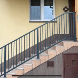 Zewnętrzne schody z szarą balustradą z pionowych prętów, wykończone płytkami imitującymi marmur, prowadzące do wejścia z brązowymi drzwiami, z widoczną drewnianą kolumną podtrzymującą zadaszenie.