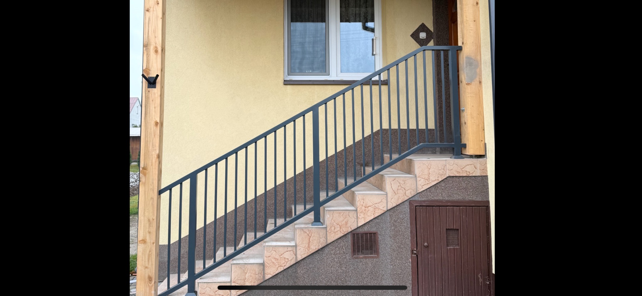 Zewnętrzne schody z szarą balustradą z pionowych prętów, wykończone płytkami imitującymi marmur, prowadzące do wejścia z brązowymi drzwiami, z widoczną drewnianą kolumną podtrzymującą zadaszenie.