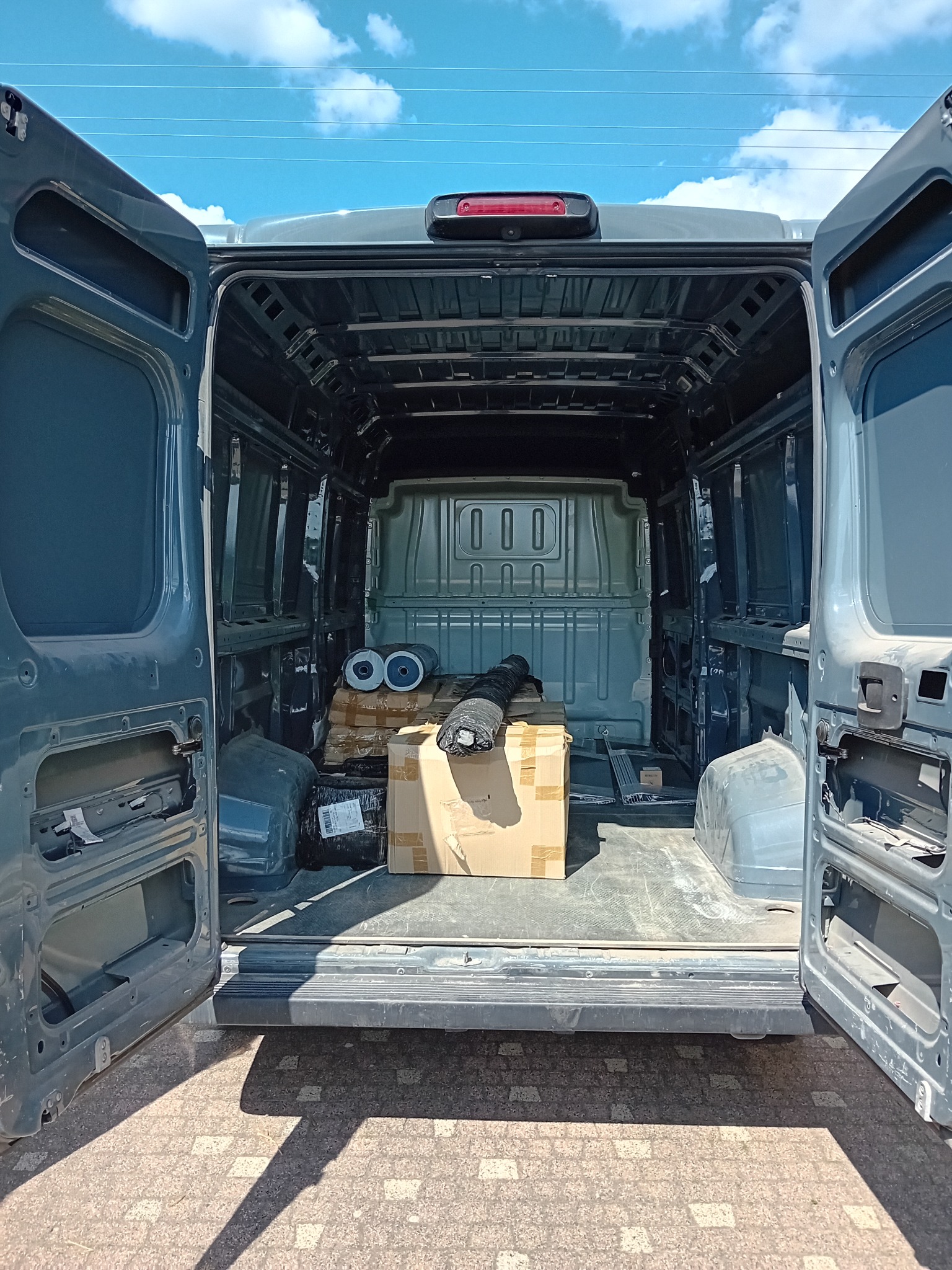 Wnętrze szarego busa z otwartymi drzwiami, widoczne kartony, rolki i pakunki przygotowane do transportu, błękitne niebo w tle.