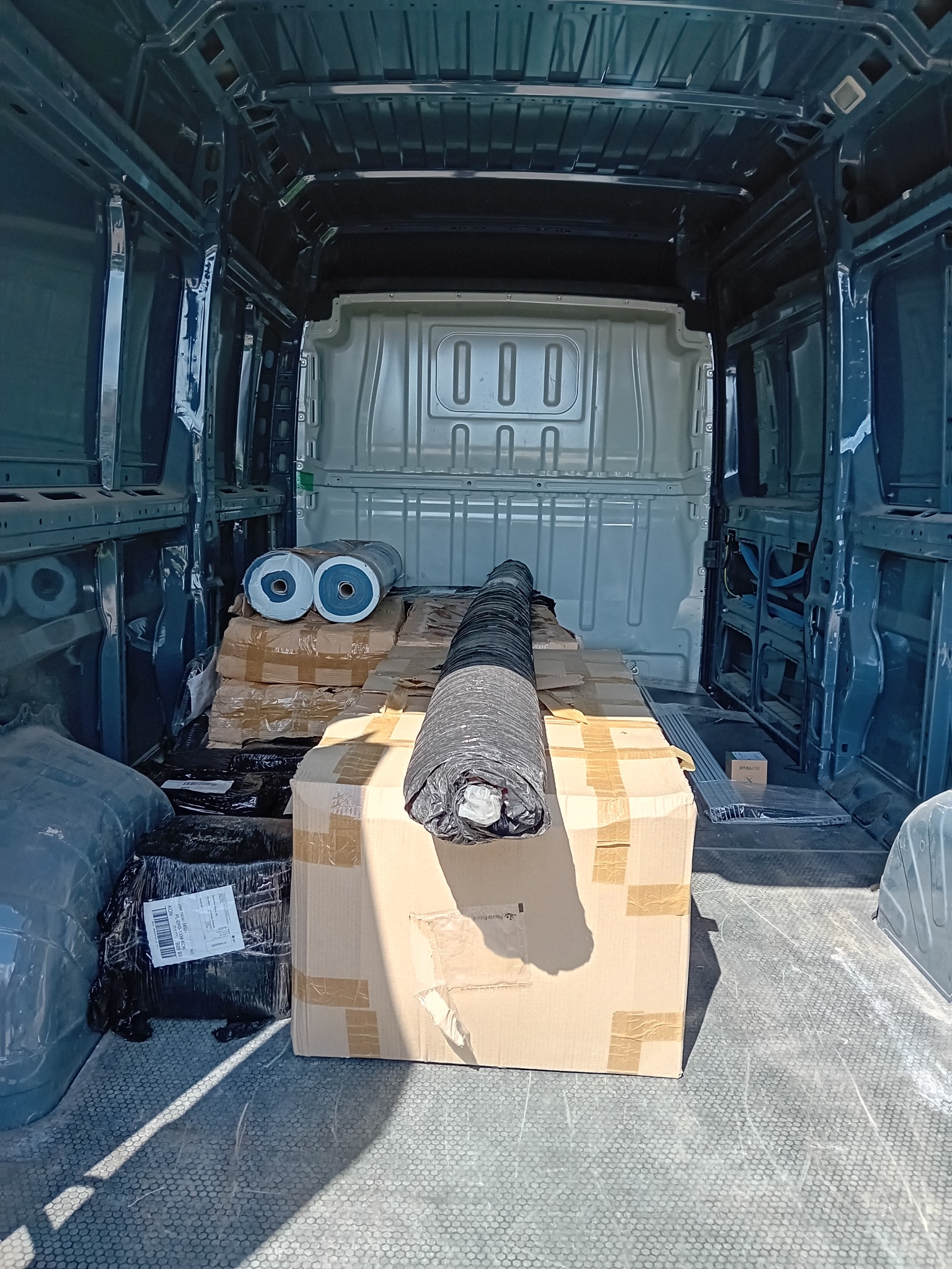 Wnętrze furgonetki załadowane kartonami, rolkami materiału zabezpieczonymi folią, gotowe do przewozu.