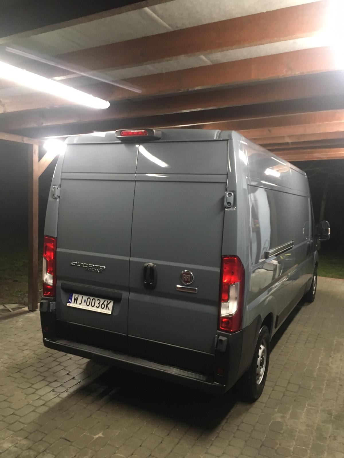 Szary samochód dostawczy Fiat Ducato zaparkowany pod zadaszeniem z drewnianymi belkami i oświetleniem jarzeniowym, widok z tyłu, tablica rejestracyjna WJ-0036K.