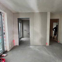 Perfect Line ARKADIUSZ MYSZAK - Wnętrze w trakcie remontu: widoczne ściany z płyt karton-gips, surowy beton na podłodze, okno z rusztowaniem na zewnątrz, pracownik w tle i oznaczenia laserowe na ścianach.