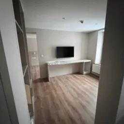 Pomalowane na beżowo wnętrze pokoju z drewnianą podłogą, białym biurkiem pod telewizorem i grzejnikiem pod oknem, widok zza regału.