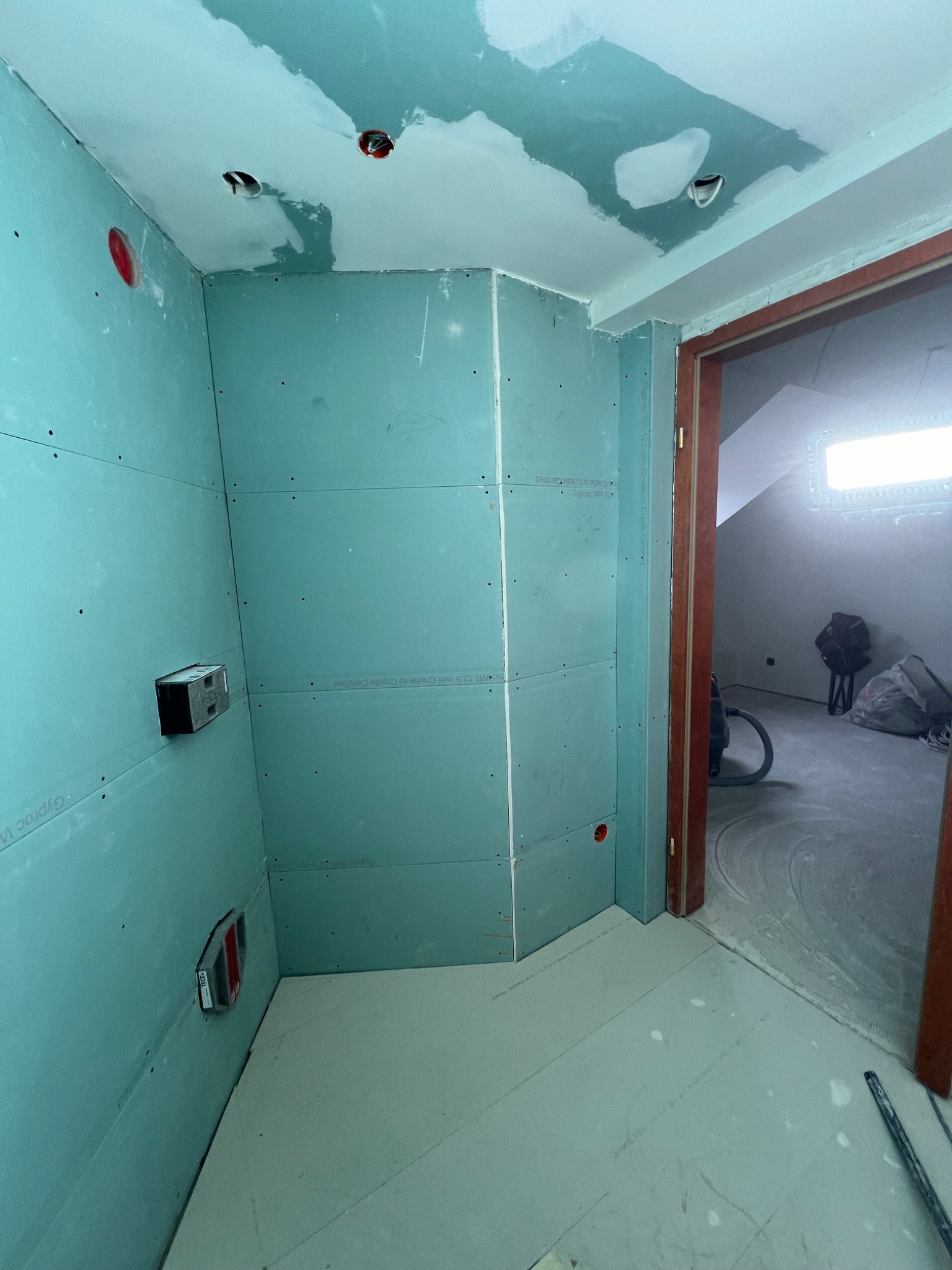 Wnętrze w trakcie montażu płyt kartonowo-gipsowych, widoczne ściany z zamontowanymi płytami, puszki elektryczne, częściowo wykończony sufit oraz otwarte drzwi do pomieszczenia z workami i odkurzaczem.