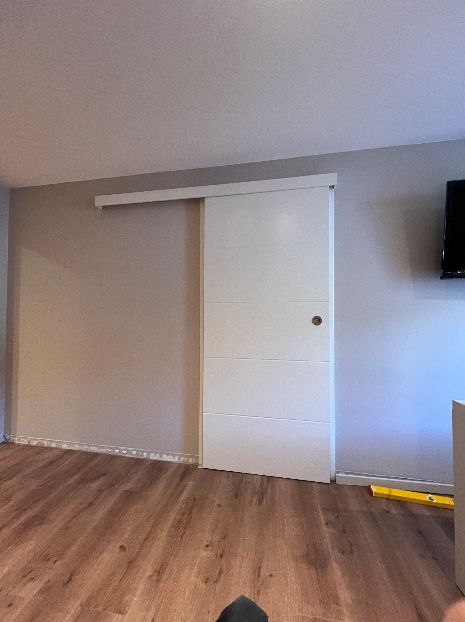 Białe, przesuwne drzwi z poziomymi panelami, zamontowane w szarej ścianie, z widoczną listwą przypodłogową w trakcie montażu i poziomnicą leżącą na podłodze z paneli.