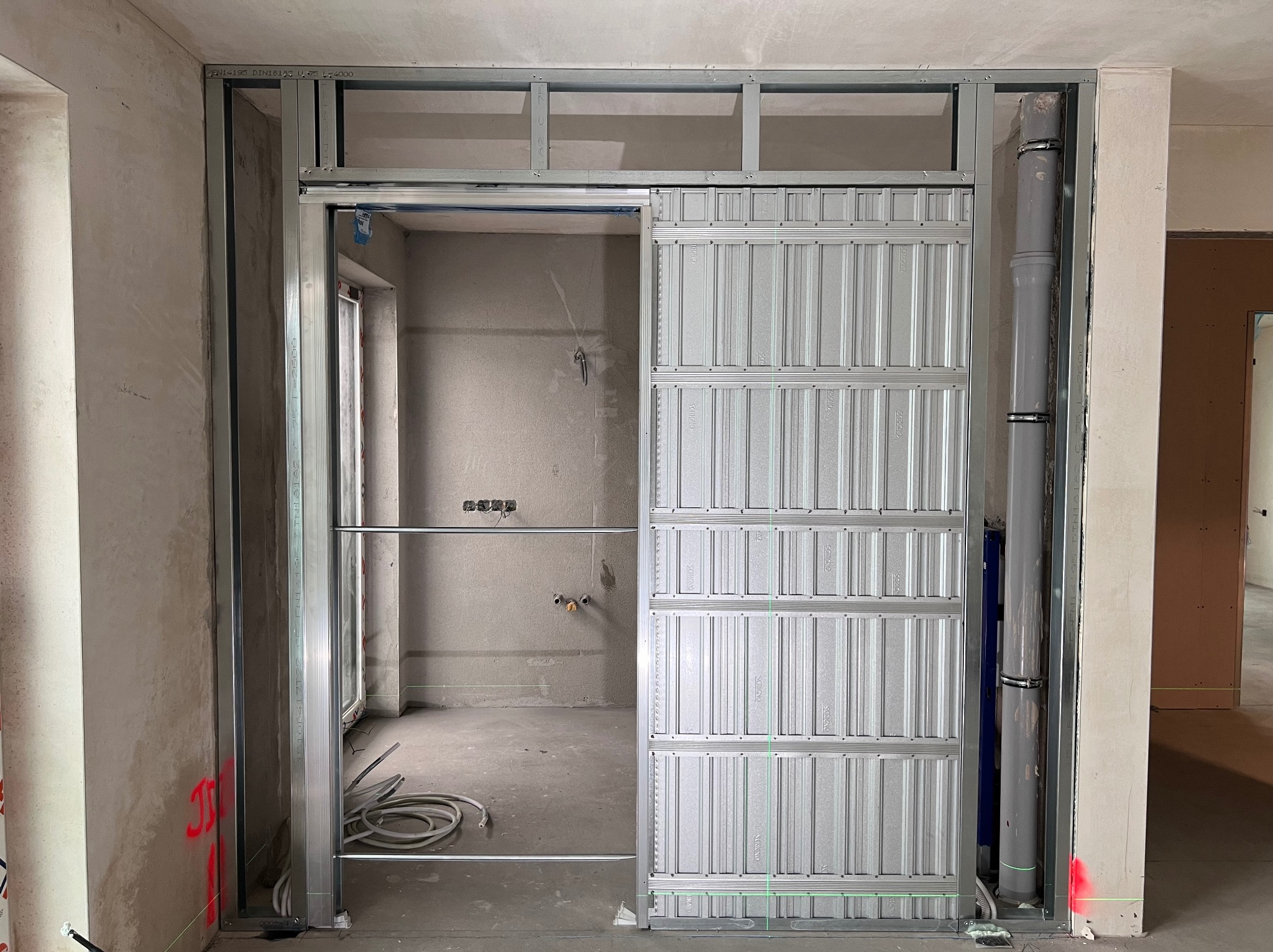 Montaż metalowej konstrukcji pod ścianę z płyt kartonowo-gipsowych, widoczna surowa ściana w tle oraz instalacja sanitarna.