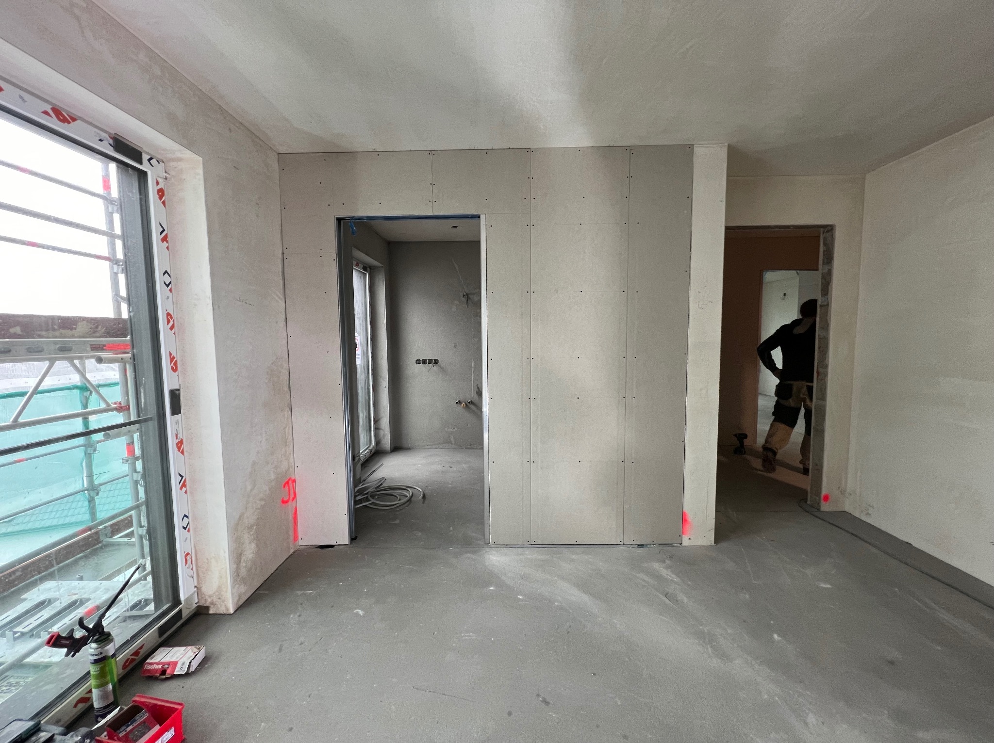 Wnętrze w trakcie remontu: widoczne ściany z płyt karton-gips, surowy beton na podłodze, okno z rusztowaniem na zewnątrz, pracownik w tle i oznaczenia laserowe na ścianach.