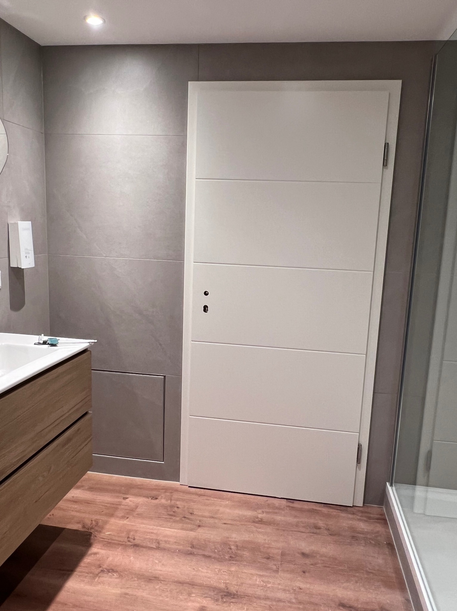 Nowoczesna łazienka z szarymi płytkami, drewnianą szafką pod umywalką i białą, panelową ścianką oddzielającą prysznic.