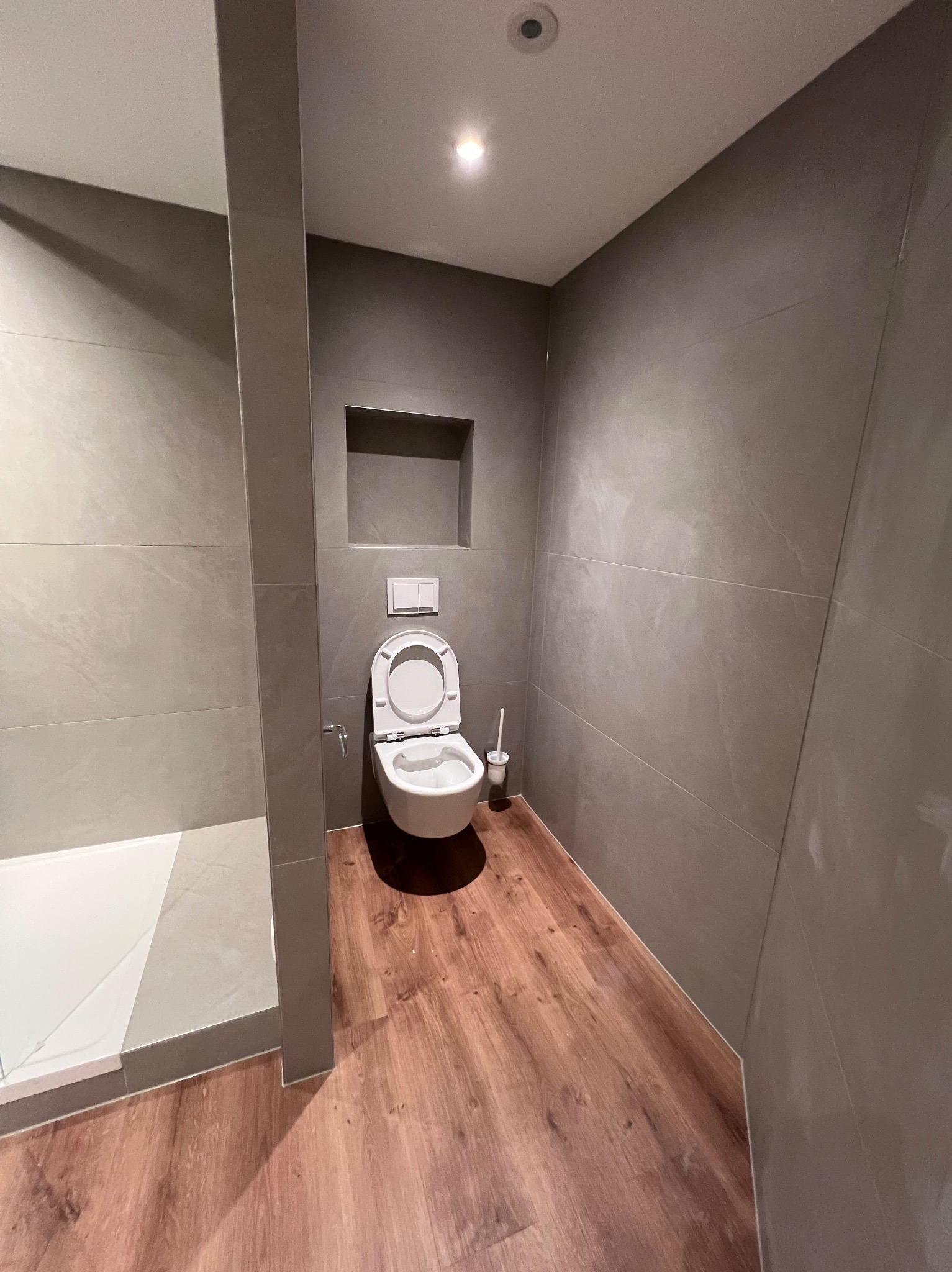 Nowoczesna łazienka z szarymi płytkami imitującymi kamień na ścianach, drewnopodobną podłogą, wiszącą toaletą z otwartą klapą i wnęką w ścianie.