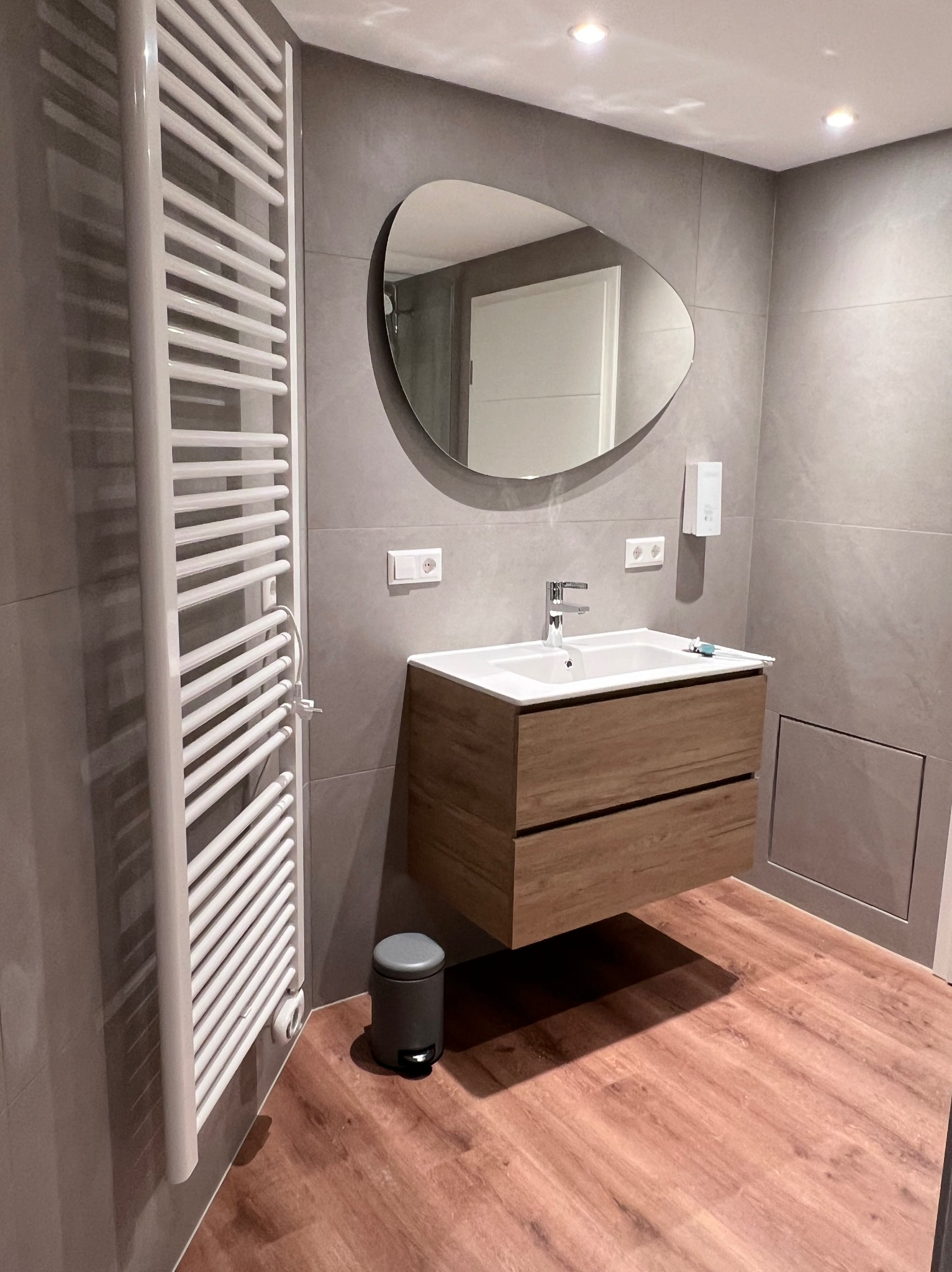 Nowoczesna łazienka z wiszącą szafką z umywalką, owalnym lustrem, grzejnikiem drabinkowym i szarą glazurą ścienną oraz podłogą imitującą drewno.