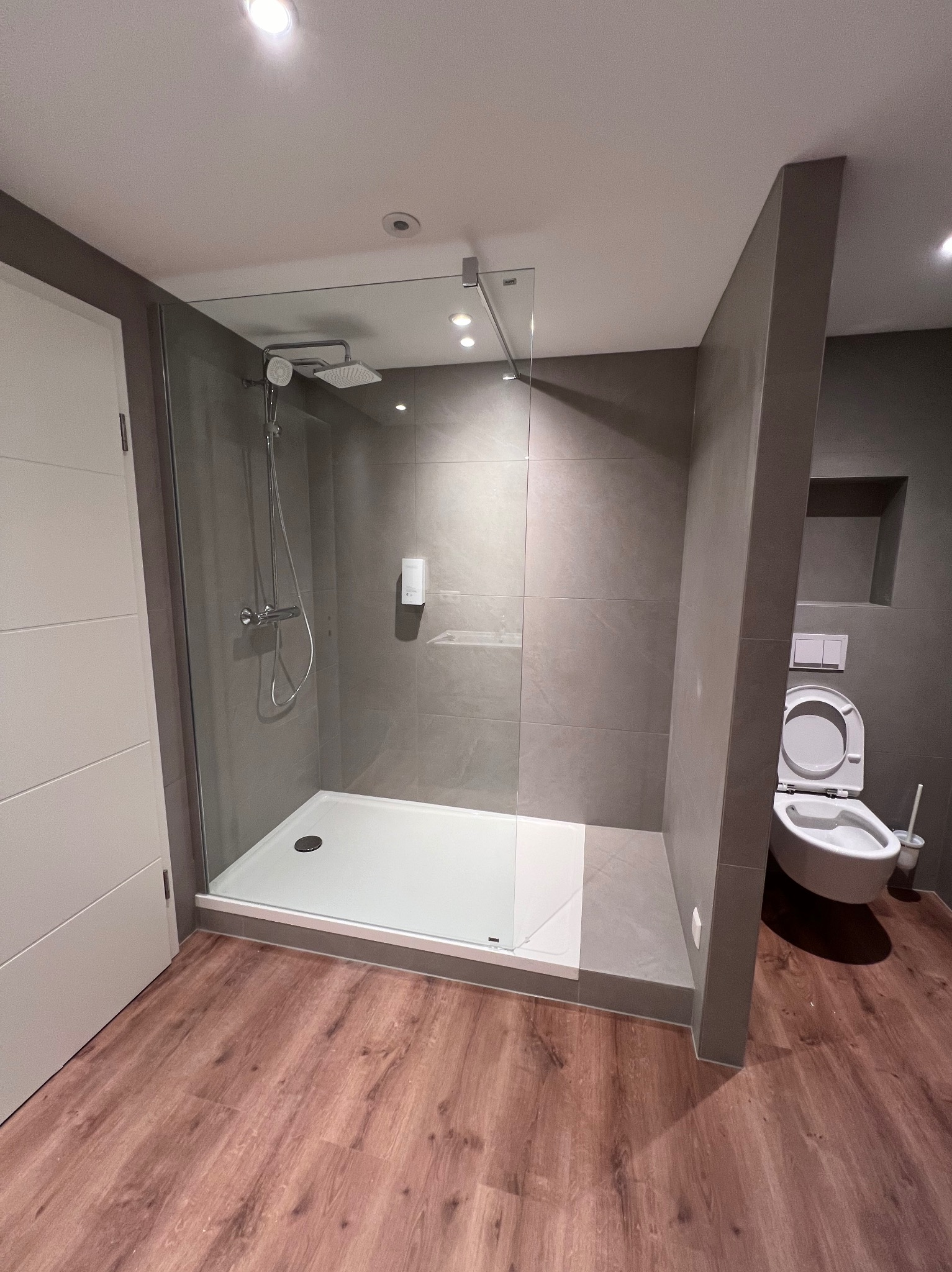 Nowoczesna łazienka z kabiną prysznicową typu walk-in, szare płytki na ścianach, drewnopodobna podłoga oraz wisząca toaleta z otwartą klapą.