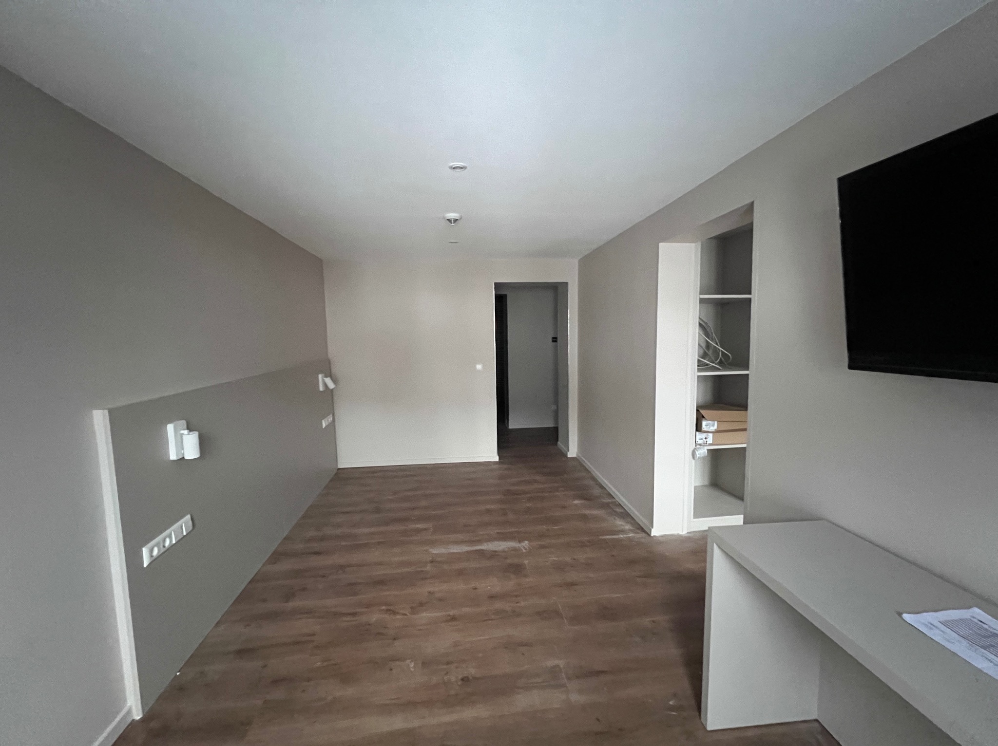 Wnętrze pokoju po remoncie z panelami podłogowymi, świeżo pomalowanymi ścianami w odcieniach szarości, widocznymi gniazdkami elektrycznymi i wnęką z półkami. Na ścianie wisi telewizor.