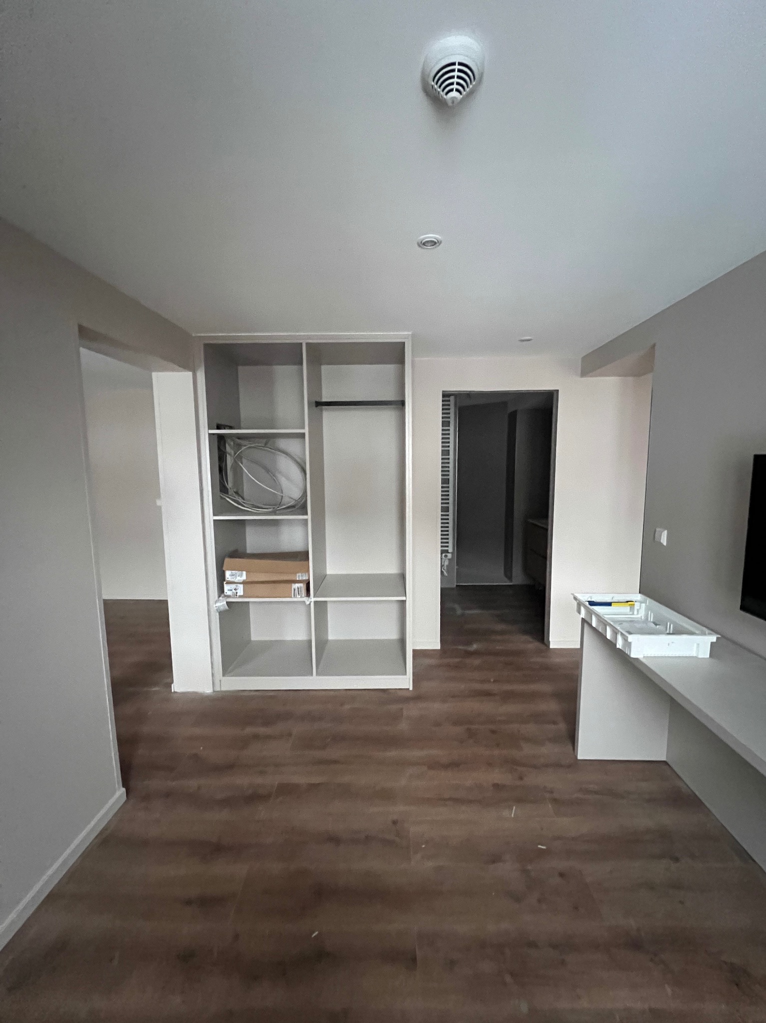 Wnętrze mieszkania w trakcie wykańczania: widoczna zabudowa szafy z półkami, częściowo przygotowana, z widocznymi przewodami i kartonami na półkach, oraz blat roboczy z pojemnikiem na narzędzia,...