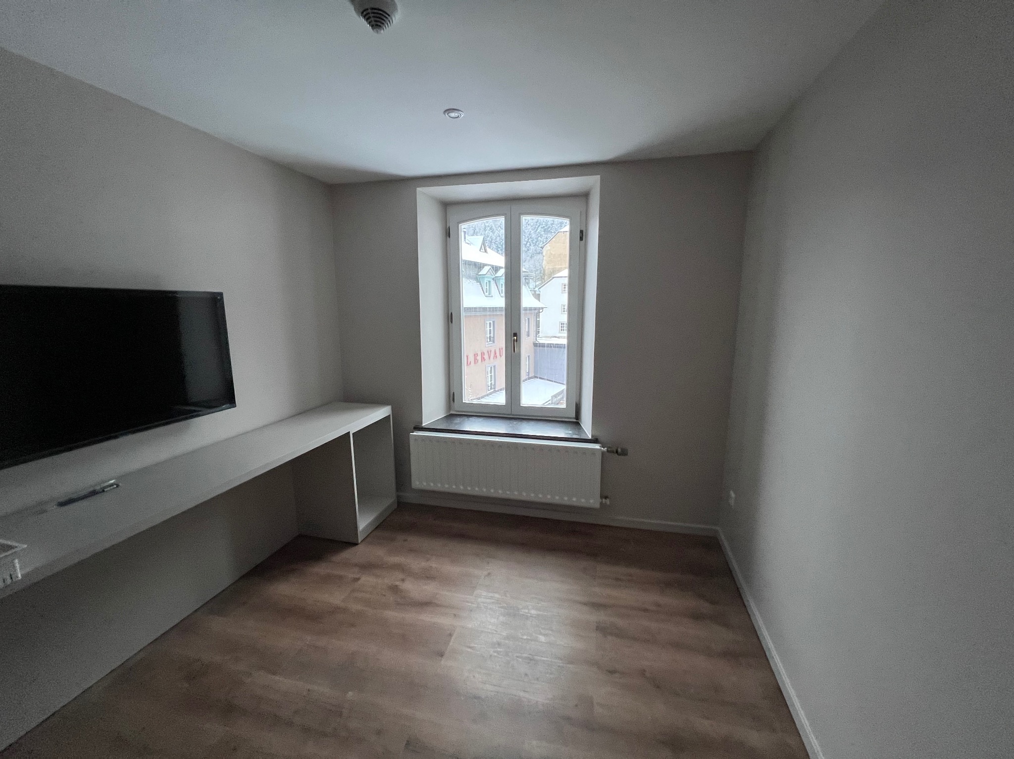 Wnętrze pokoju po remoncie z drewnopodobną podłogą, jasnymi ścianami i oknem z widokiem na zabudowania miejskie w zimowej scenerii. Pod oknem grzejnik, z lewej strony zamontowany długi blat...