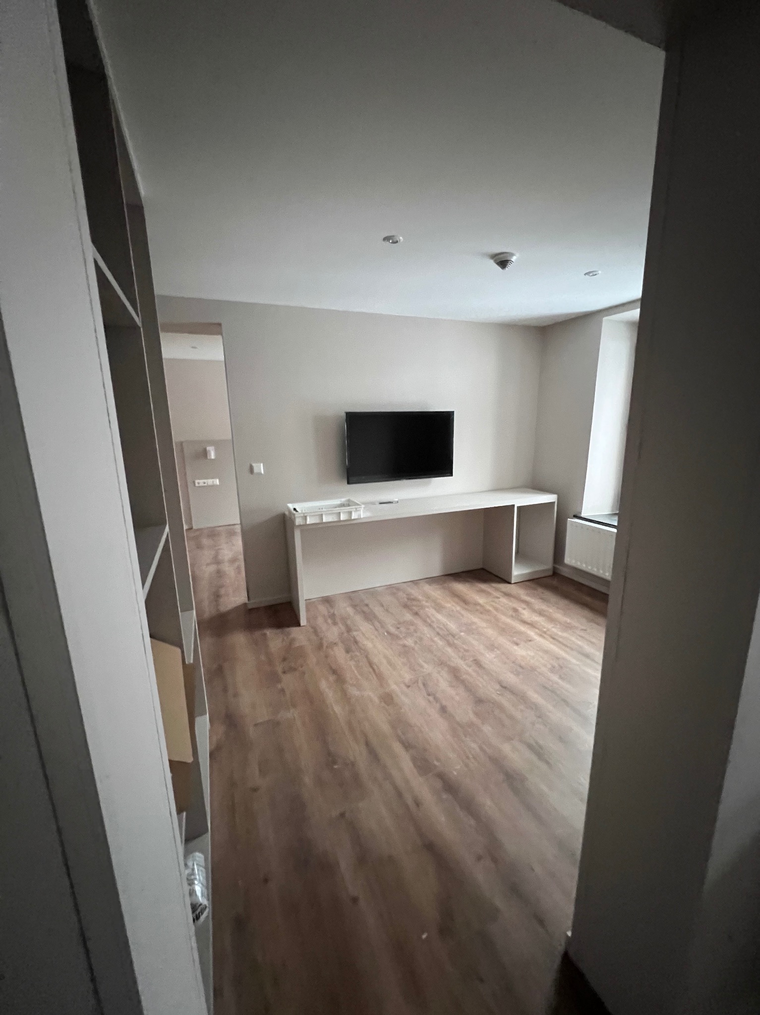 Pomalowane na beżowo wnętrze pokoju z drewnianą podłogą, białym biurkiem pod telewizorem i grzejnikiem pod oknem, widok zza regału.