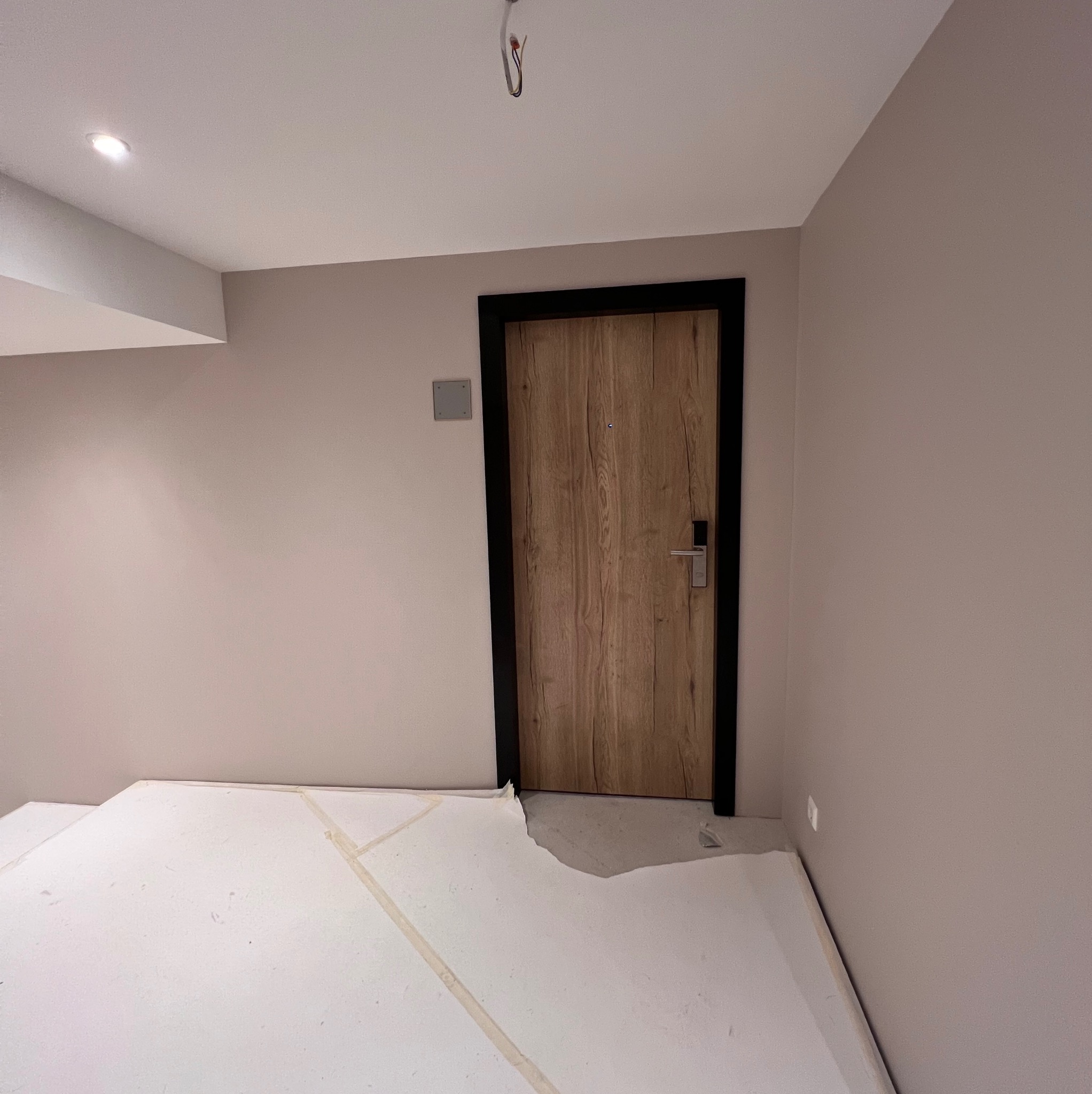 Pokój w trakcie remontu z zabezpieczoną podłogą folią malarską, drzwi wejściowe z czarną ościeżnicą i drewnianym panelem, ściany pomalowane na jasny kolor.