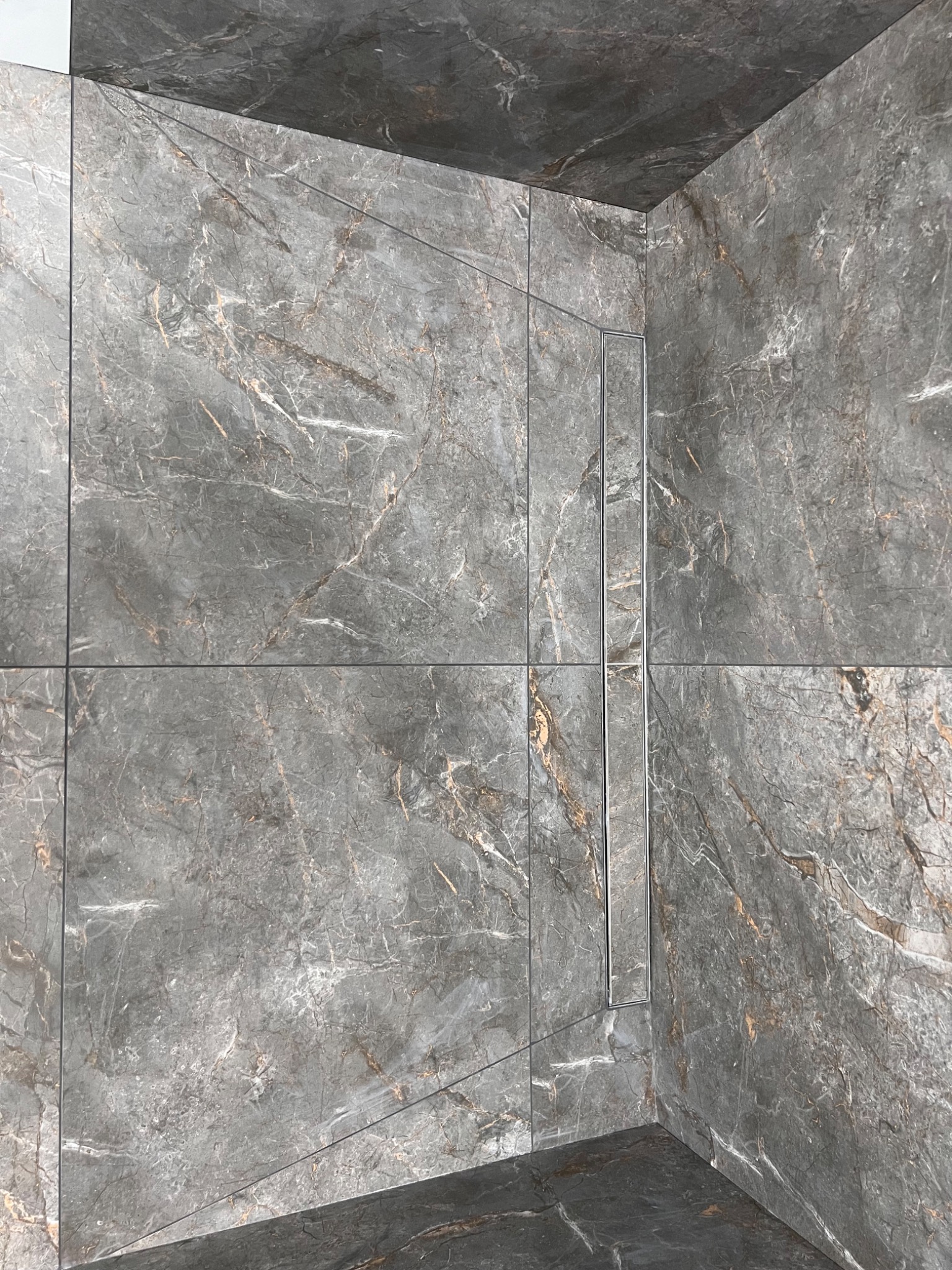 Wykończona kabina prysznicowa z szarymi, marmurkowymi płytkami i chromowanym odpływem liniowym. Estetyczne połączenie płytek na ścianach i podłodze.