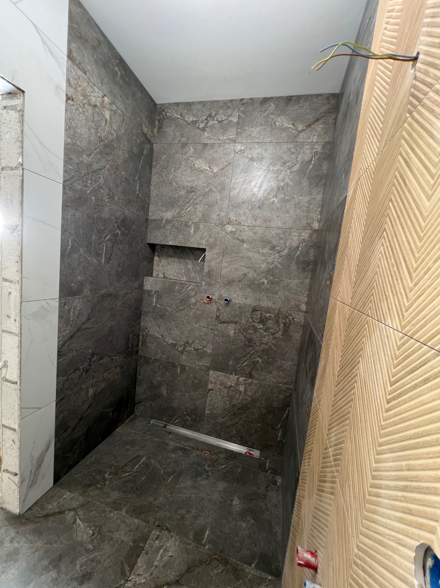 Wykończona kabina prysznicowa z szarymi płytkami imitującymi marmur i drewnopodobną ścianą z wzorem jodełki. Widoczny odpływ liniowy i wnęka w ścianie.