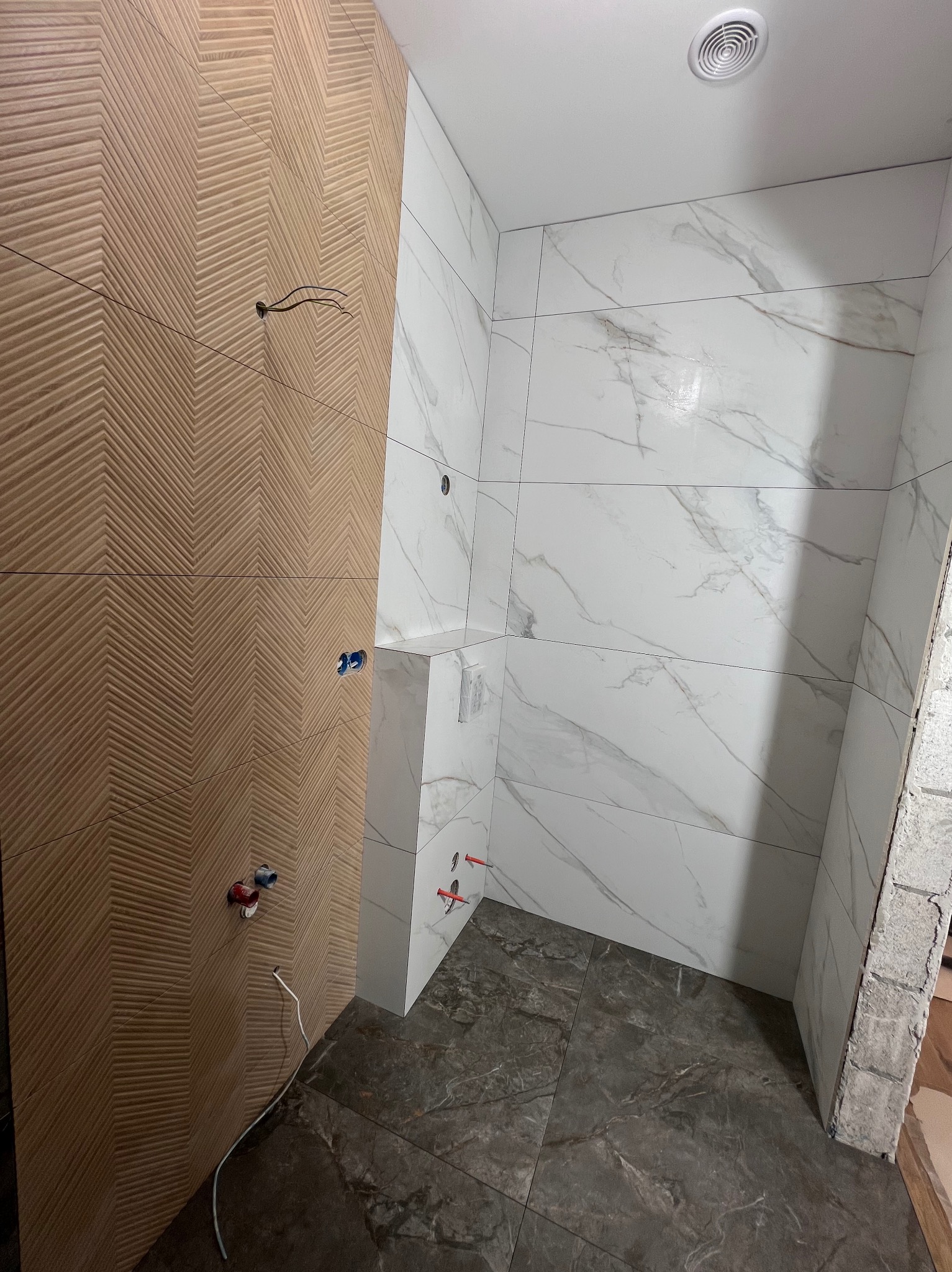 Wykończona łazienka z drewnopodobną ścianą w jodełkę i białymi płytkami imitującymi marmur. Widoczne wyprowadzenia instalacji wodnej i elektrycznej.