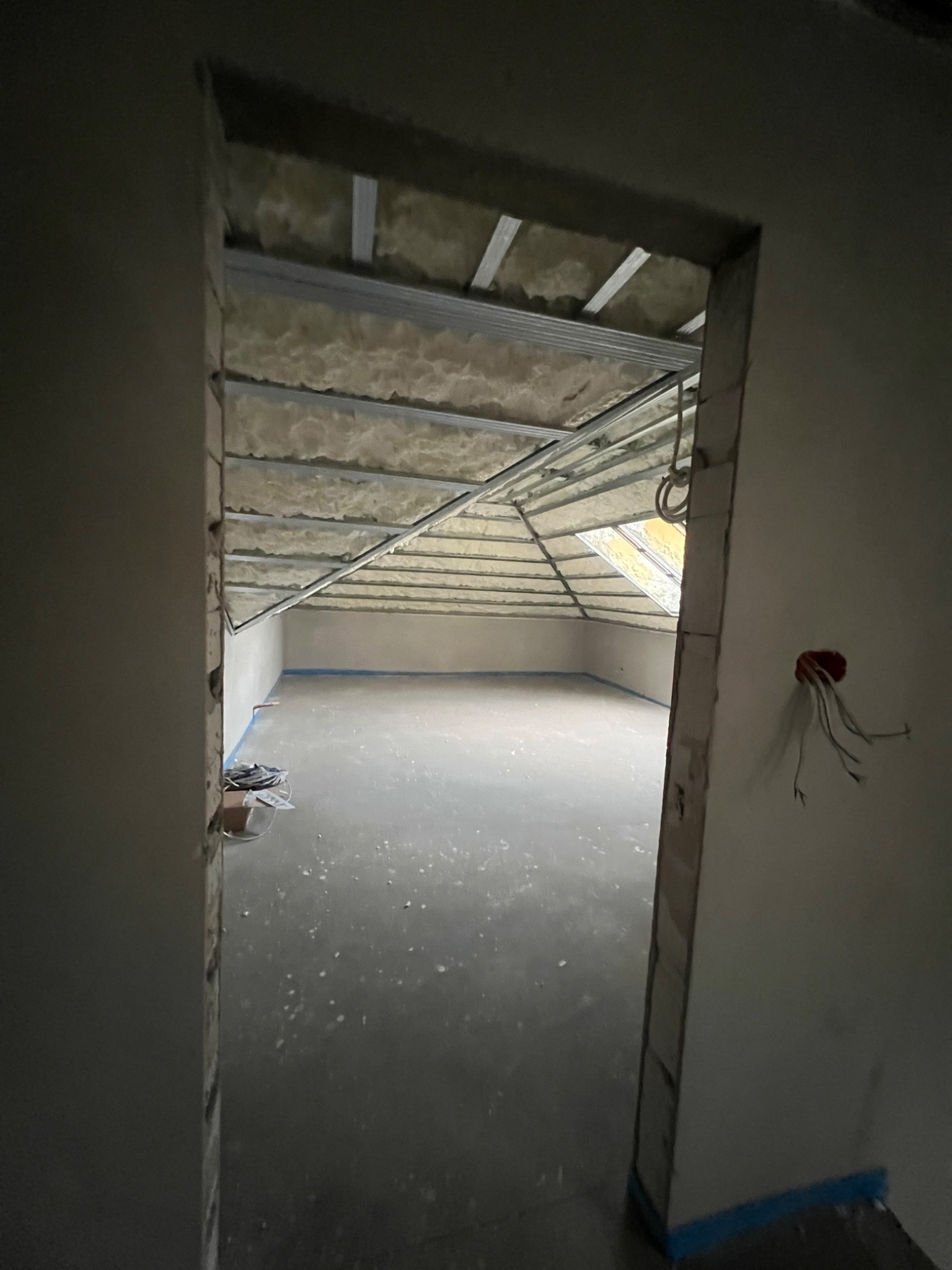 Poddasze w trakcie adaptacji: widok przez surowy otwór drzwiowy na ocieplony strop z metalową konstrukcją i okno dachowe. W tle przygotowana posadzka.