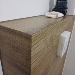 ŁOPATKA - Blat szafki łazienkowej z drewnopodobnym wykończeniem, na którym leży robocza lampka akumulatorowa i ściereczka, obok widoczna kostka do fugowania, tło stanowią białe płytki.