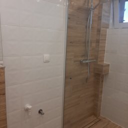 ŁOPATKA - Nowoczesna kabina prysznicowa walk-in z odpływem liniowym, wykończona białymi płytkami strukturalnymi i panelami imitującymi drewno, wyposażona w deszczownicę i słuchawkę prysznicową.