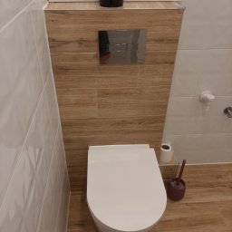 ŁOPATKA - Nowoczesna toaleta wisząca z chromowanym przyciskiem spłukującym, obudowana płytami imitującymi drewno, uzupełniona o białe płytki ścienne i akcesoria łazienkowe.