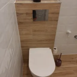 Nowoczesna toaleta wisząca z chromowanym przyciskiem spłukującym, obudowana płytami imitującymi drewno, uzupełniona o białe płytki ścienne i akcesoria łazienkowe.