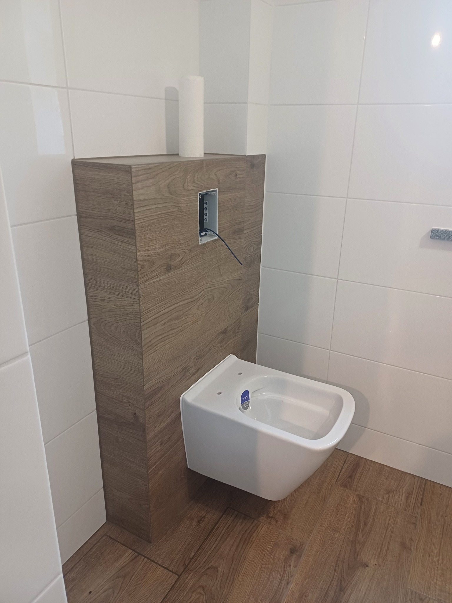 Wisząca toaleta z zabudowaną spłuczką w obudowie imitującej drewno, białe płytki na ścianach i drewnopodobna podłoga.