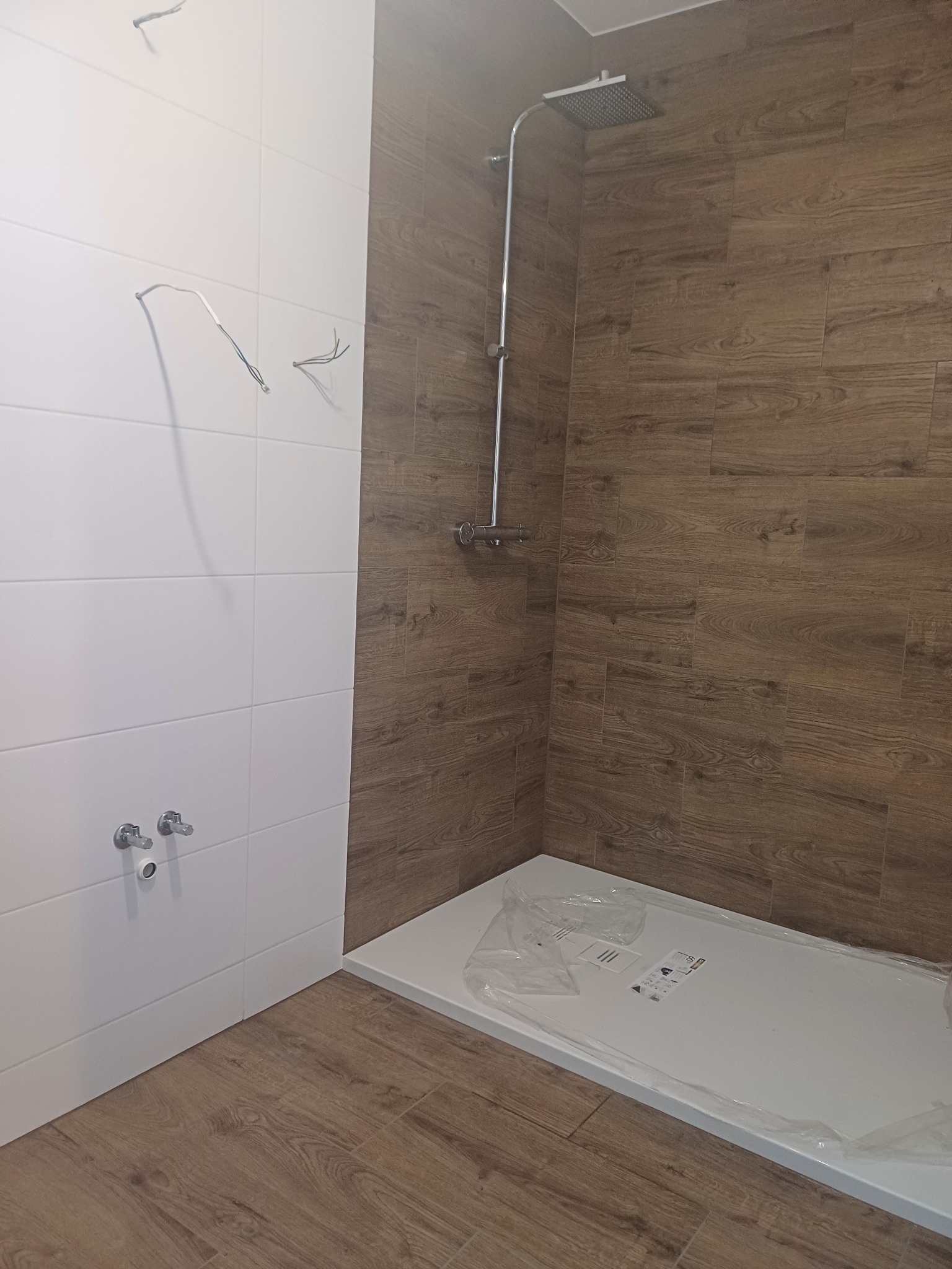 Niedokończona łazienka z białą płytką na ścianie, wystającymi przewodami elektrycznymi, brązową płytką imitującą drewno na ścianie prysznicowej i brodzikiem w folii ochronnej.