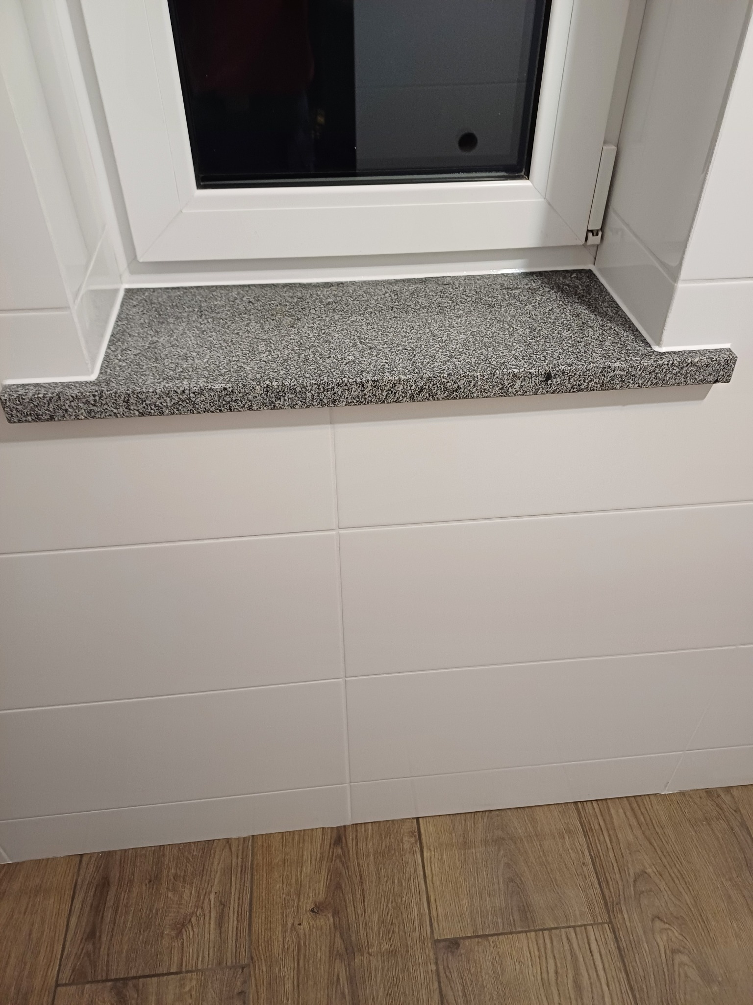 Szary, nakrapiany parapet z konglomeratu pod oknem w łazience z białymi, prostokątnymi płytkami i drewnopodobną podłogą.