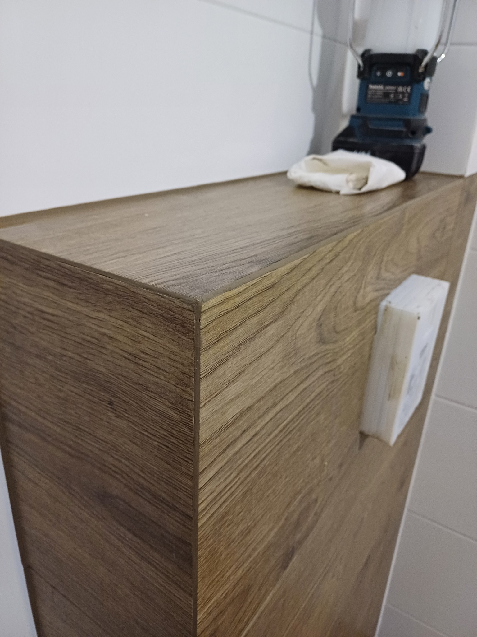 Blat szafki łazienkowej z drewnopodobnym wykończeniem, na którym leży robocza lampka akumulatorowa i ściereczka, obok widoczna kostka do fugowania, tło stanowią białe płytki.