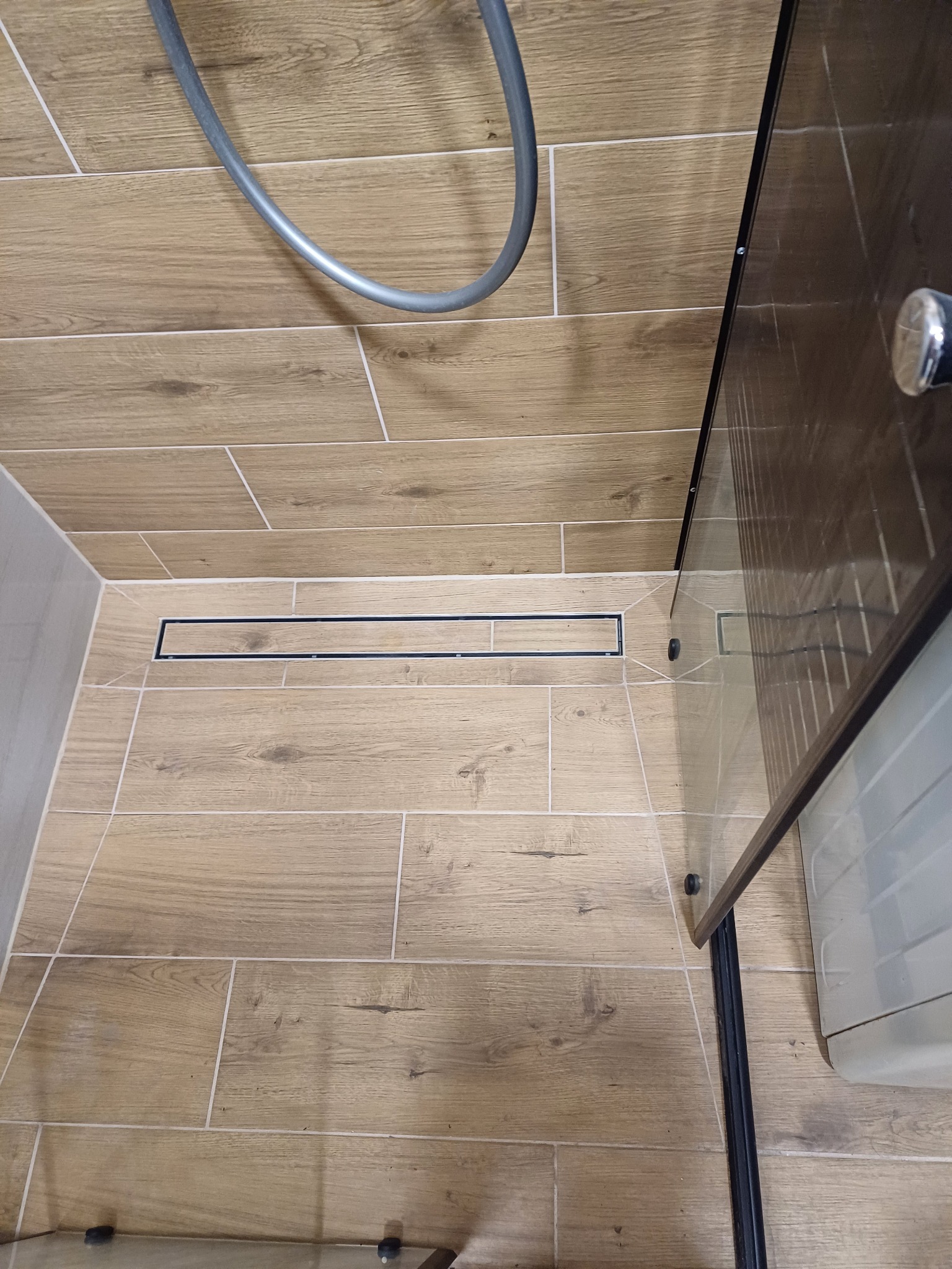 Ułożone płytki podłogowe imitujące drewno w kabinie prysznicowej z odpływem liniowym i częściowo widoczną szklaną ścianką kabiny.