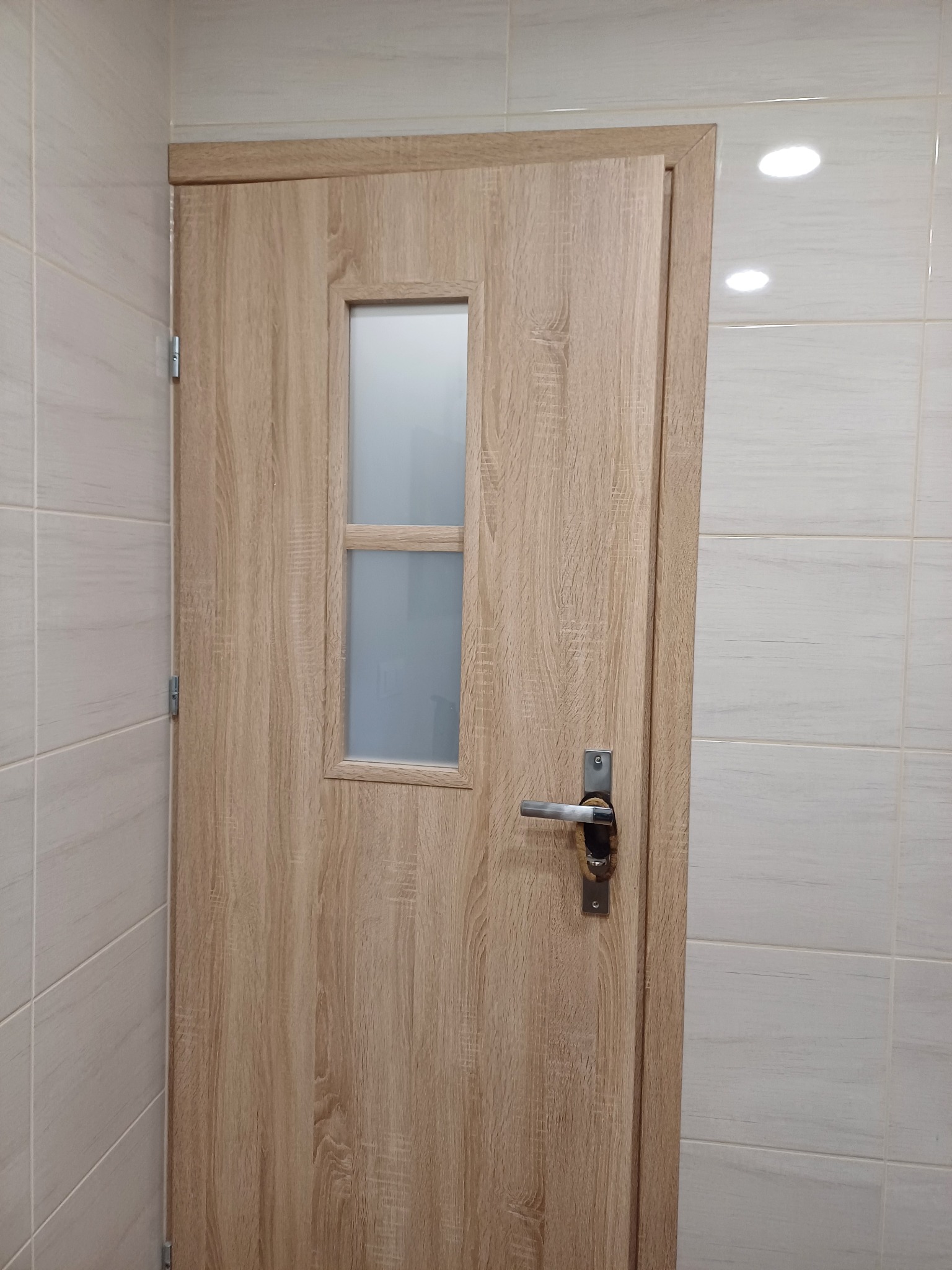 Drzwi łazienkowe w odcieniach drewna z małym, matowym przeszkleniem, zamontowane w łazience wykończonej jasnymi, kwadratowymi płytkami ceramicznymi.