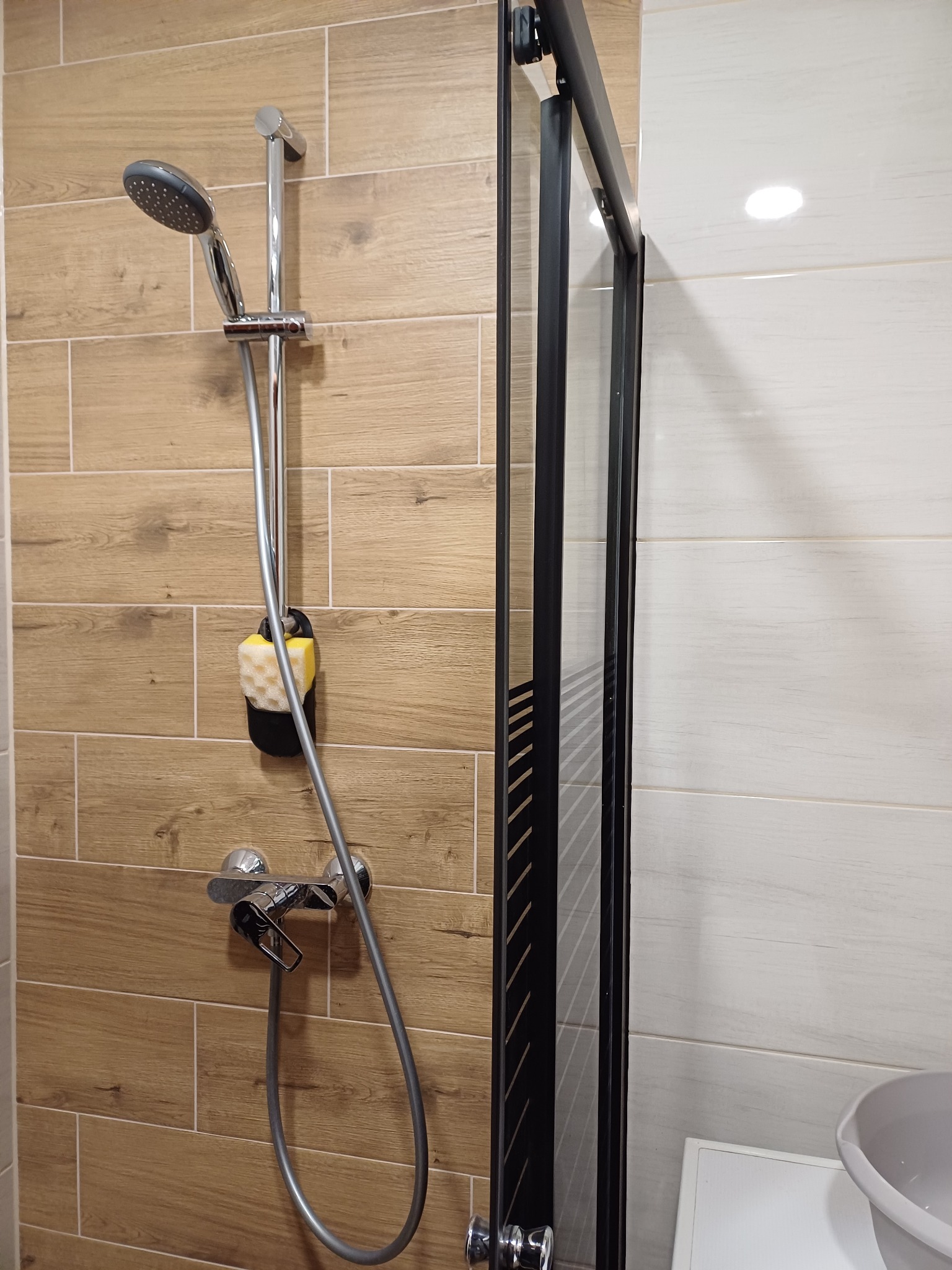 Nowoczesna kabina prysznicowa z czarną ramą, chromowaną baterią i słuchawką prysznicową na tle płytek imitujących drewno i jasnych płytek, z żółtą gąbką wiszącą na uchwycie.