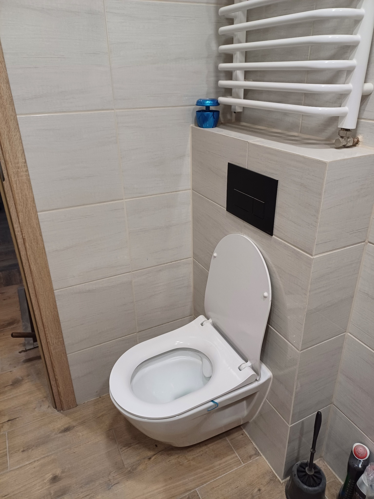 Wisząca toaleta z deską podniesioną do góry, czarny przycisk spłuczki na ścianie wyłożonej płytkami imitującymi drewno, grzejnik drabinkowy i drewnopodobna podłoga w wykończonej łazience.